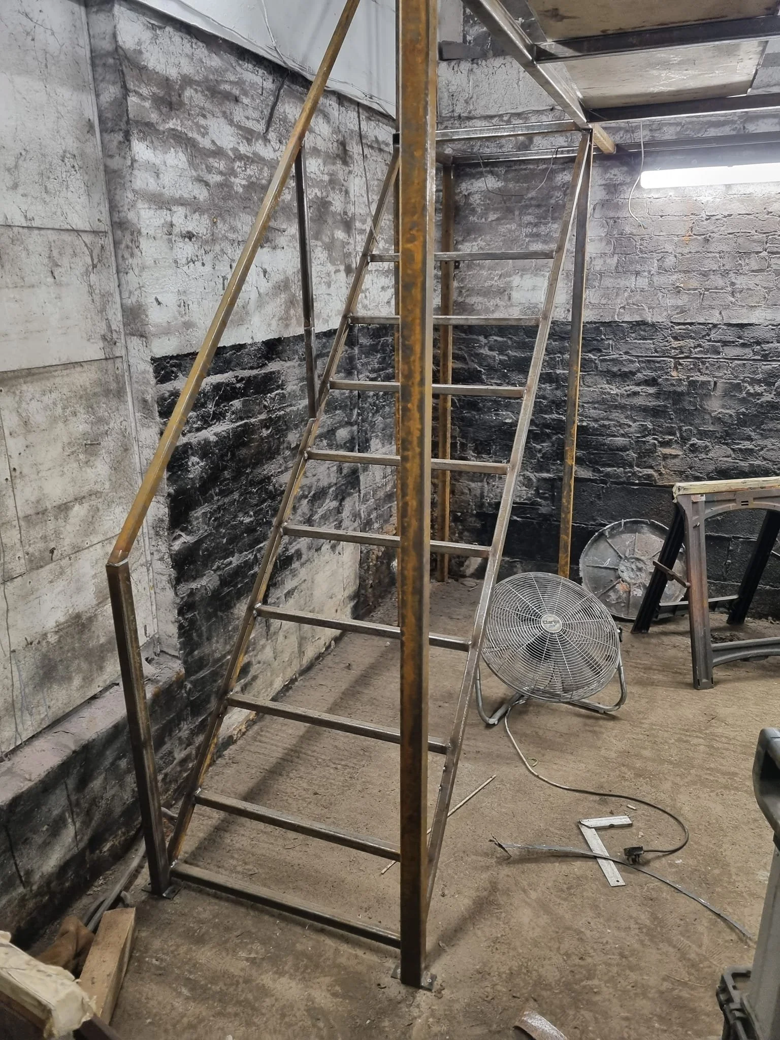 Metal scaffold with a ladder in a partially renovated basement with brick walls, a fan on the floor, and construction tools nearby.
