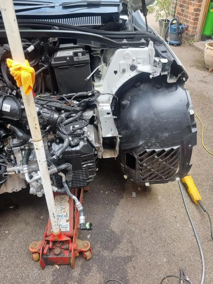 Close-up of a car with the front bumper removed, exposing the engine and components, supported by orange and yellow tools in a driveway.