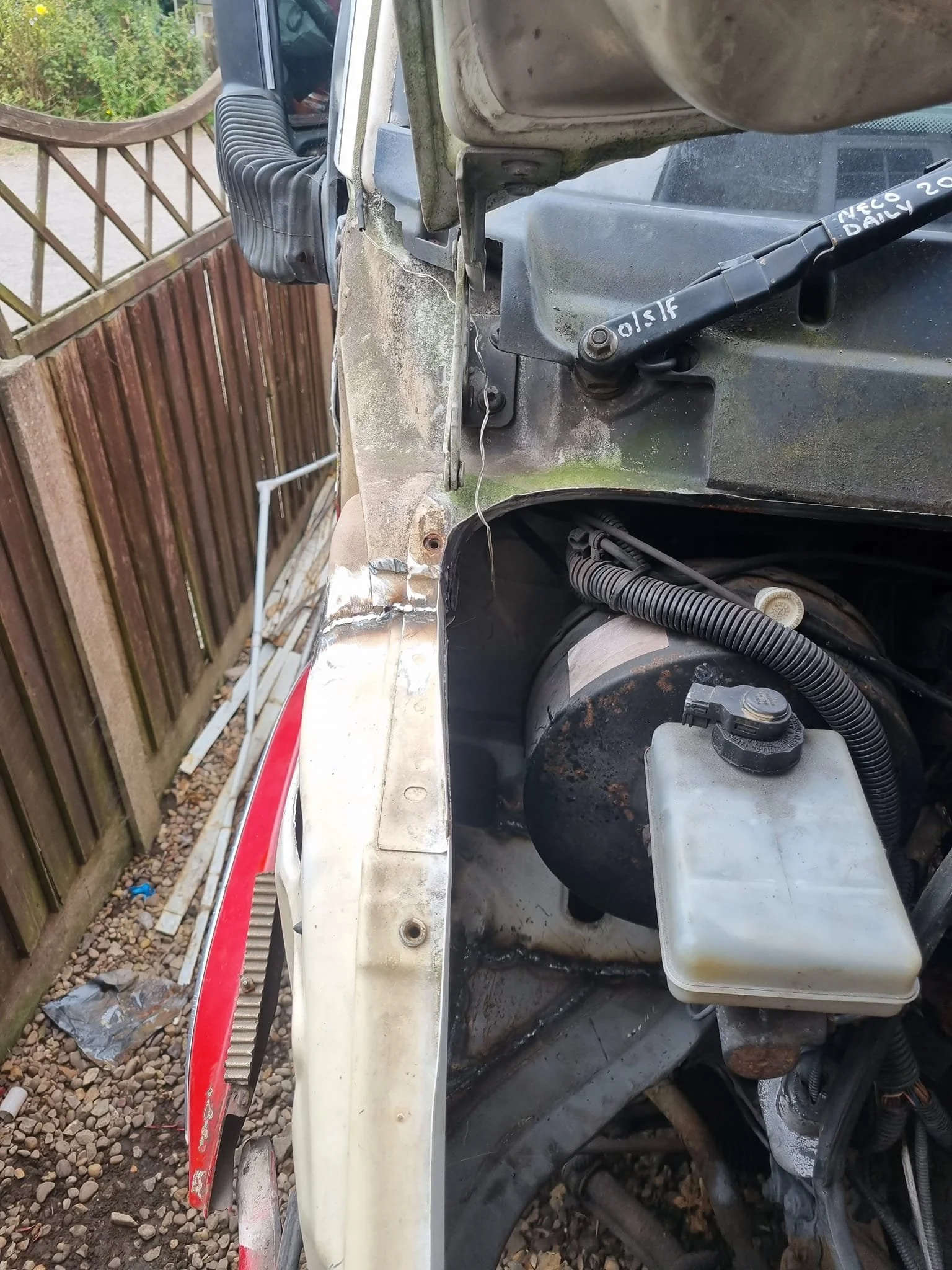 Close-up of a damaged vehicle engine bay showing rusted parts, wires, and a white fluid reservoir.