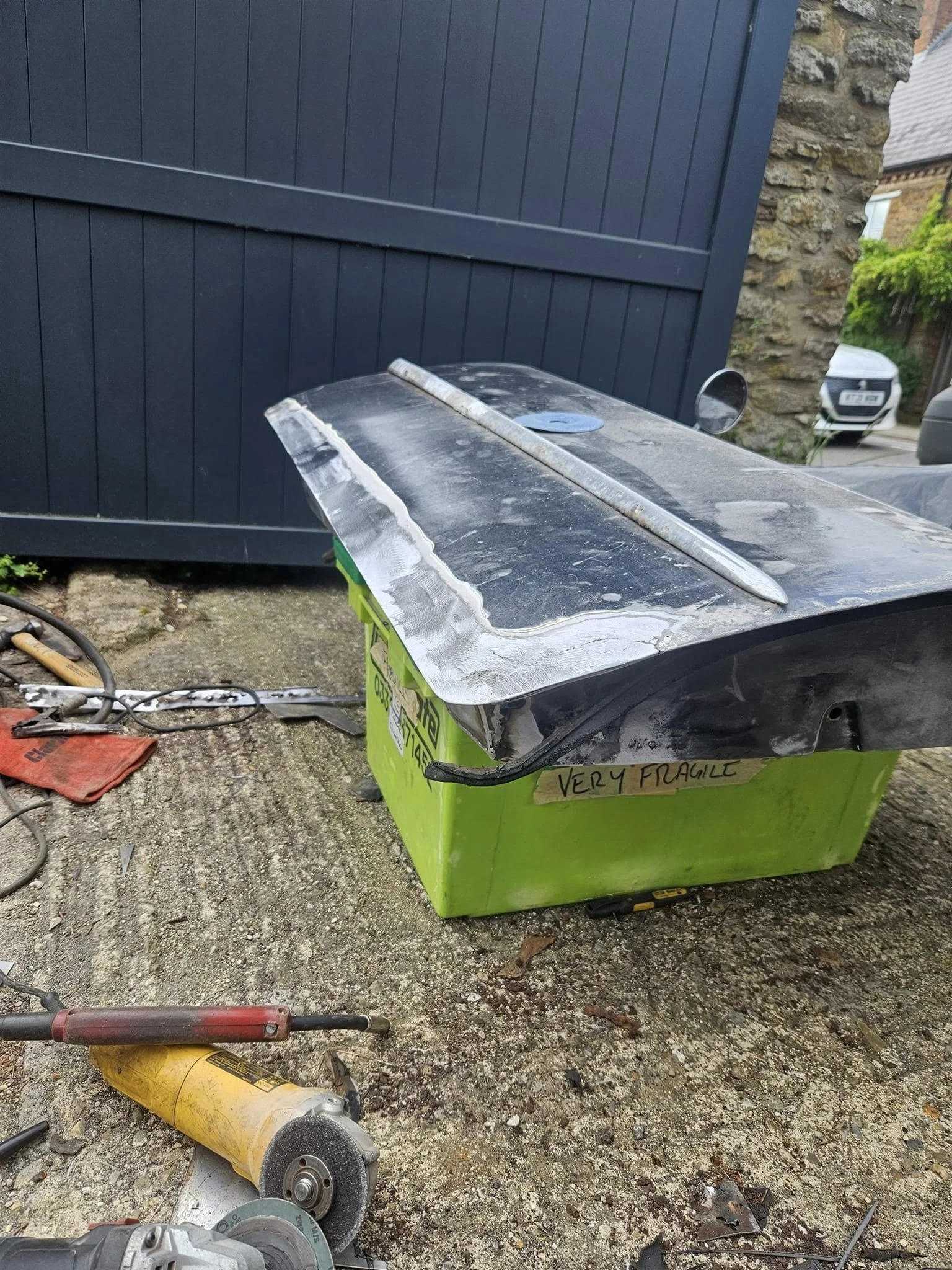 A damaged, crushed green cardboard box labeled 'Very Fragile' next to a worktable with power tools on a dirt ground outside.