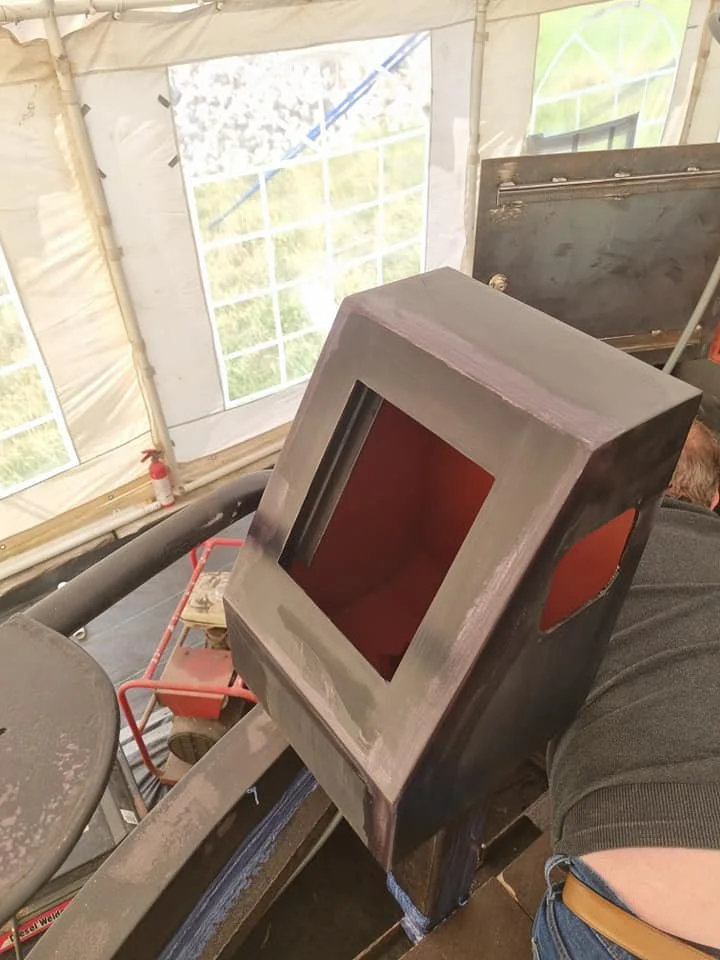 A metal enclosure with a large central opening, partially painted, resting on a work surface in a workshop or construction area.