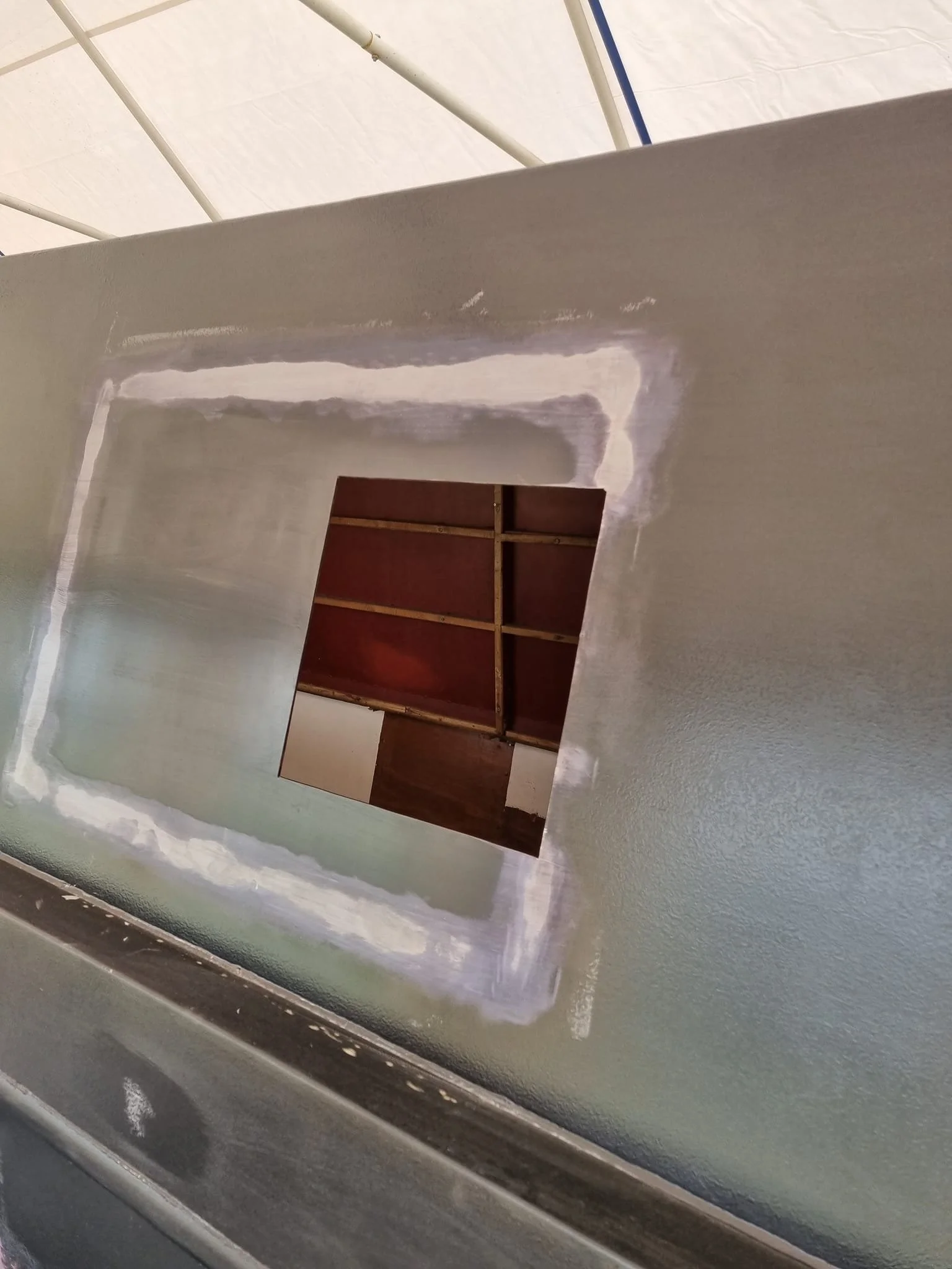 A square hole cut into a gray wall with fresh drywall joint compound around the edges, revealing a ceiling with wooden support beams in the background.