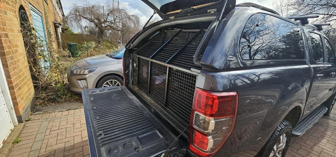 A black pickup truck with a secure cage in the truck bed parked in a driveway, with a gray SUV blocking part of the view and a brick house on the left. The truck's tailgate is open.