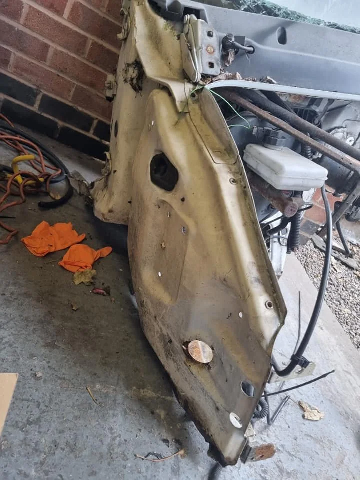 A disassembled or damaged front section of a vehicle, including the wheel well and part of the engine compartment, resting on a concrete floor with some orange cloth and tools nearby.
