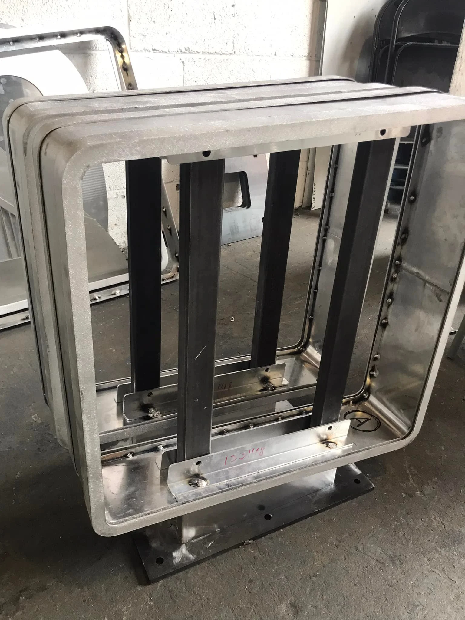 An industrial metal frame structure with welded joints, resting on a concrete floor in a workshop or warehouse setting.