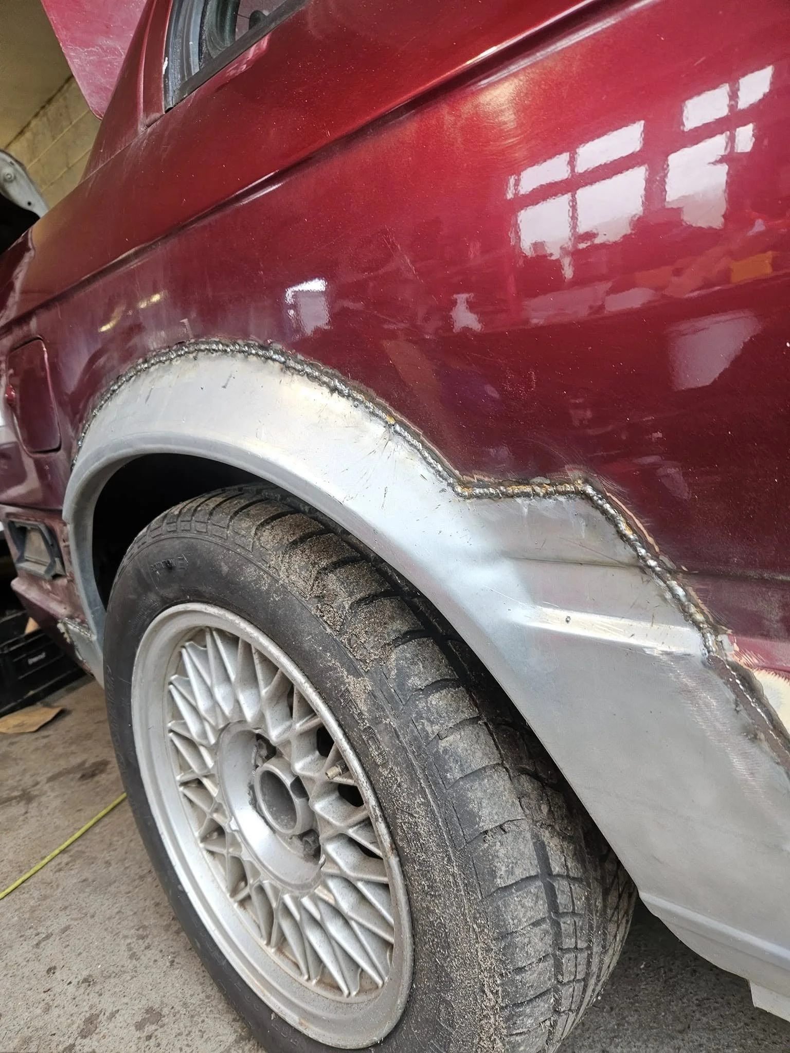 Close-up of a red car with a partially repaired and unpainted fender, showing welds and a dusty tire.