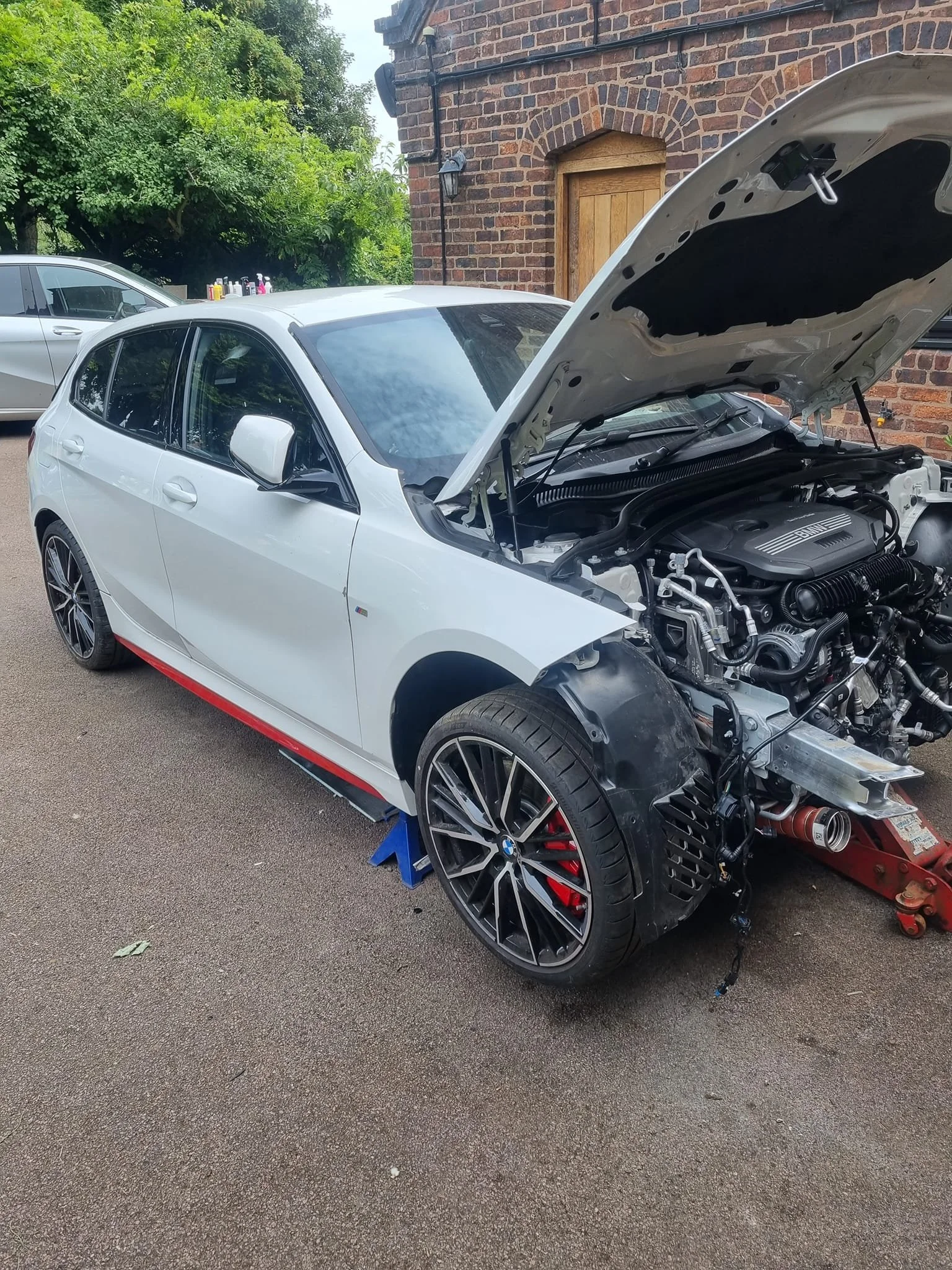 A white BMW hatchback with its hood open, supported by a jack on the front right side, parked on a driveway next to a brick house. The front right wheel is removed, exposing the brake caliper and rotor, and the car is undergoing repair or maintenance