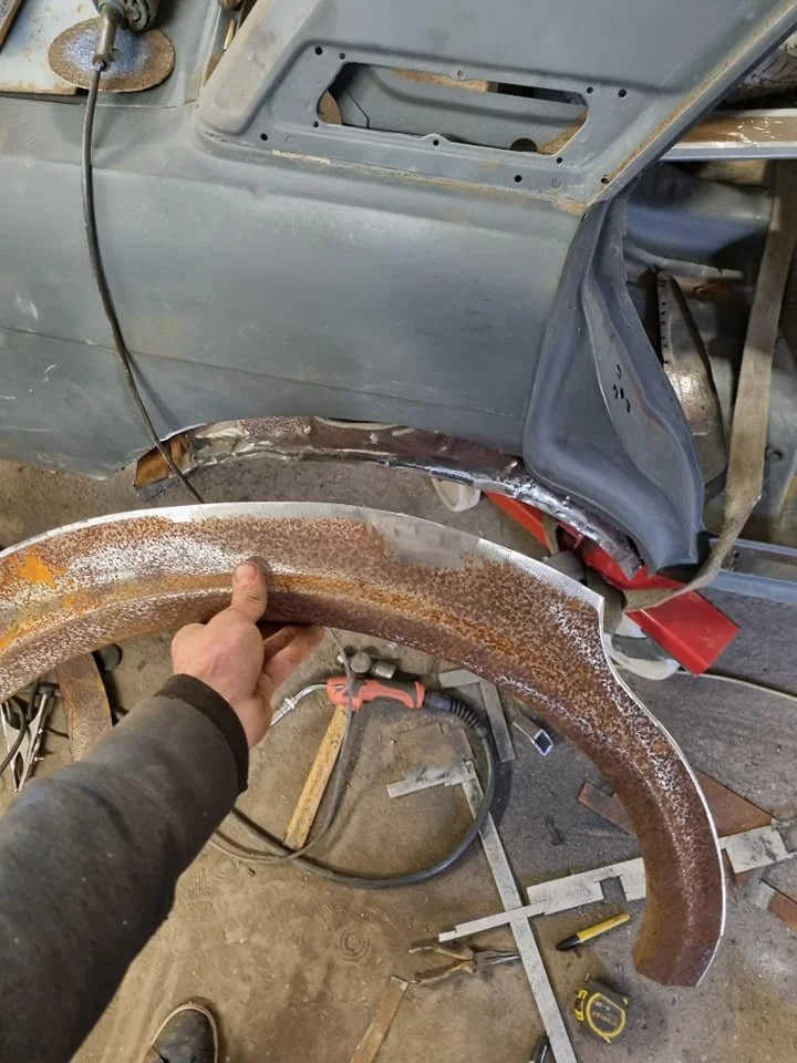 A person holding a rusty aftermarket fender near a vintage car that is being restored. The car is missing its fender and front wheel, with visible bodywork and parts in the background.