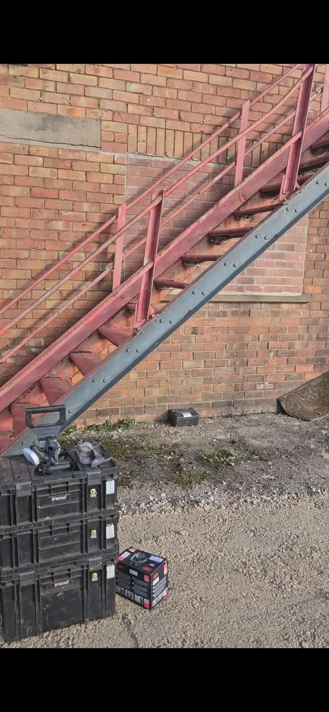 Outdoor metal fire escape stairs attached to a brick building with tools and storage boxes at the base.