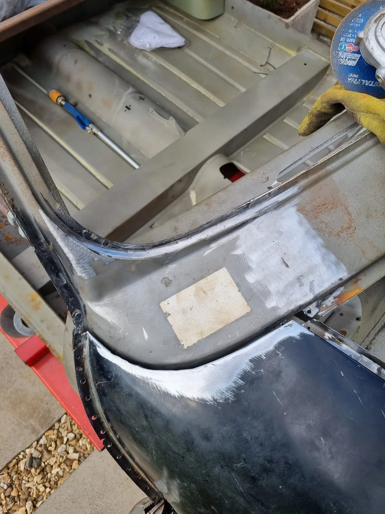 Close-up of a car repair with bodywork sanding, showing a metal surface with primer and black paint, and a workspace with tools in the background.