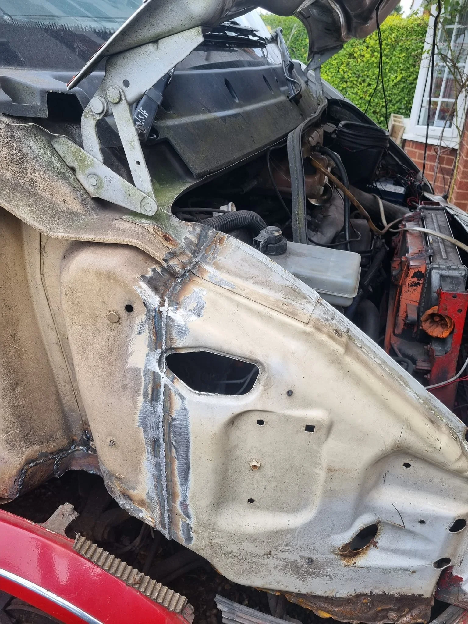 Close-up of a vintage car's front fender and engine bay with visible repair welds and rust, with an open hood and house in the background.
