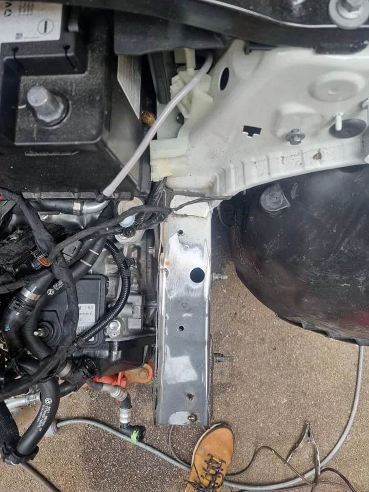 Close-up of the engine bay of a vehicle, showing engine components, wiring, and a metal support frame, with a person's foot in a work boot visible at the bottom.