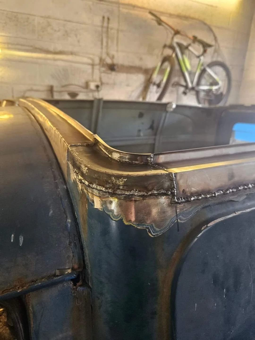 Close-up view of a metal welding joint on a black metal structure with a bicycle mounted on the wall in the background.