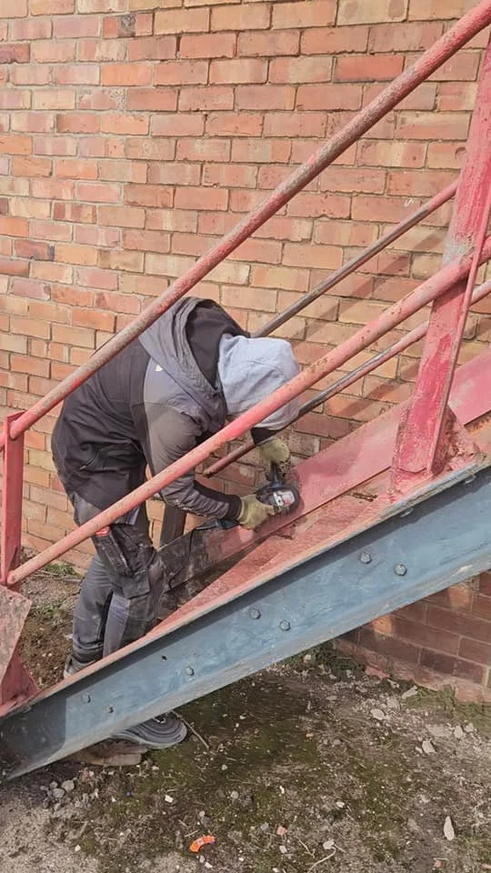 A man wearing a hoodie and work gloves is using a power drill on a metal staircase railing next to a brick wall.