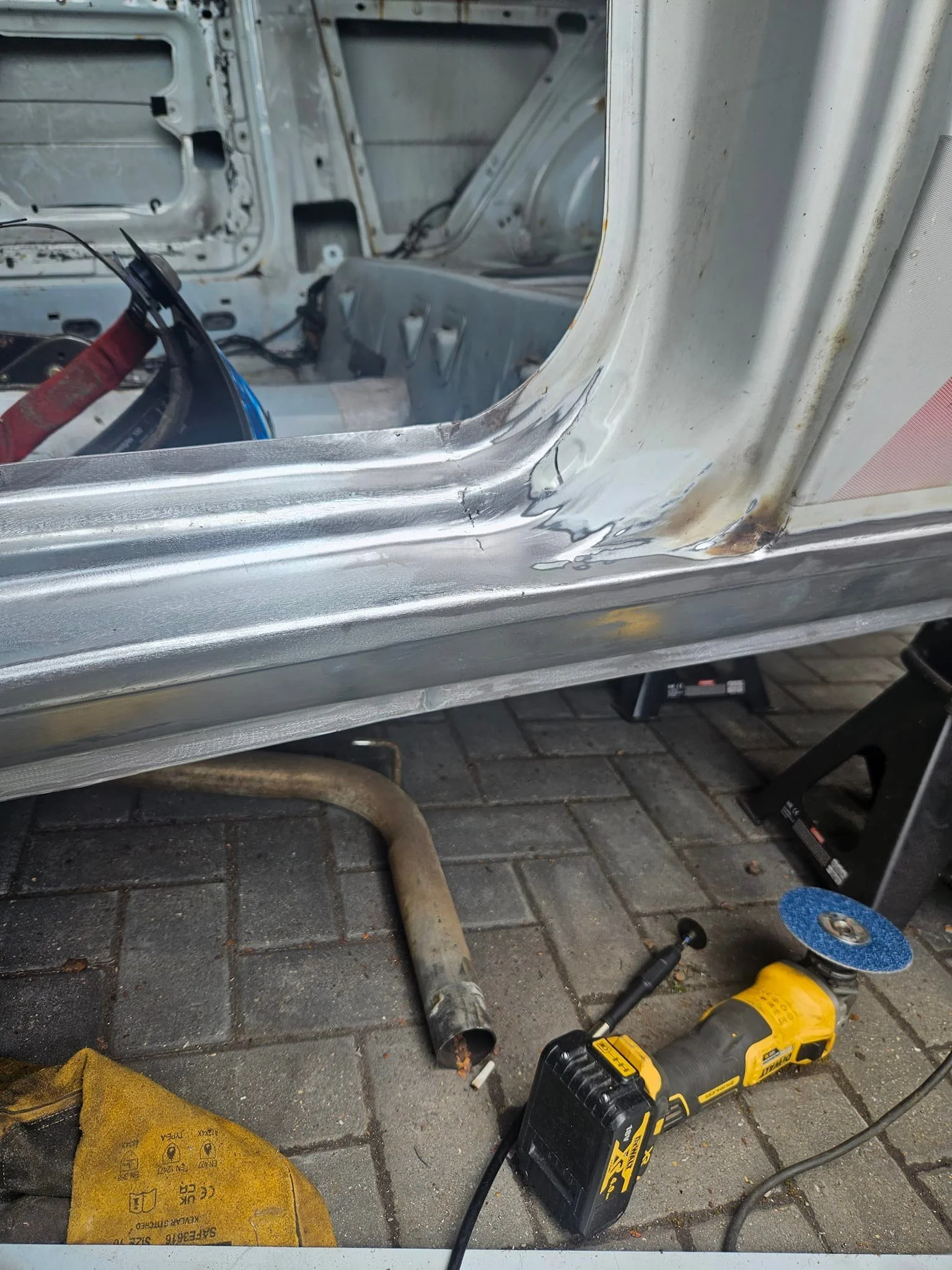 Close-up of a car's bodywork being repaired with visible metal and welding marks, along with a grinder tool on the ground nearby, on a paved surface.