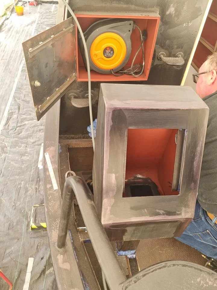 A person working on a metal project, with a partially assembled metal box or frame on a worktable. There are visible tools and a large, yellow-orange electrical cord reel inside the box.