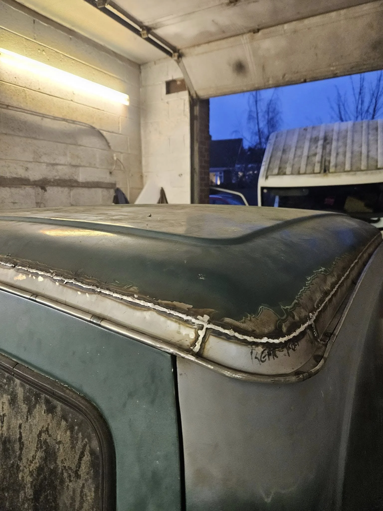 Close-up of an old, weathered vehicle roof with rust, dents, and chipped paint in a garage with a brick wall and a garage door open showing a van outside.
