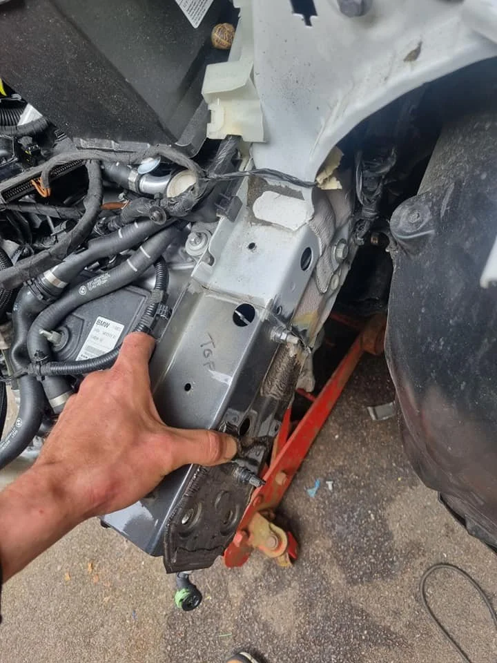 Close-up of a person's hand installing or removing a metal component in a car's engine bay, with various hoses and parts visible.