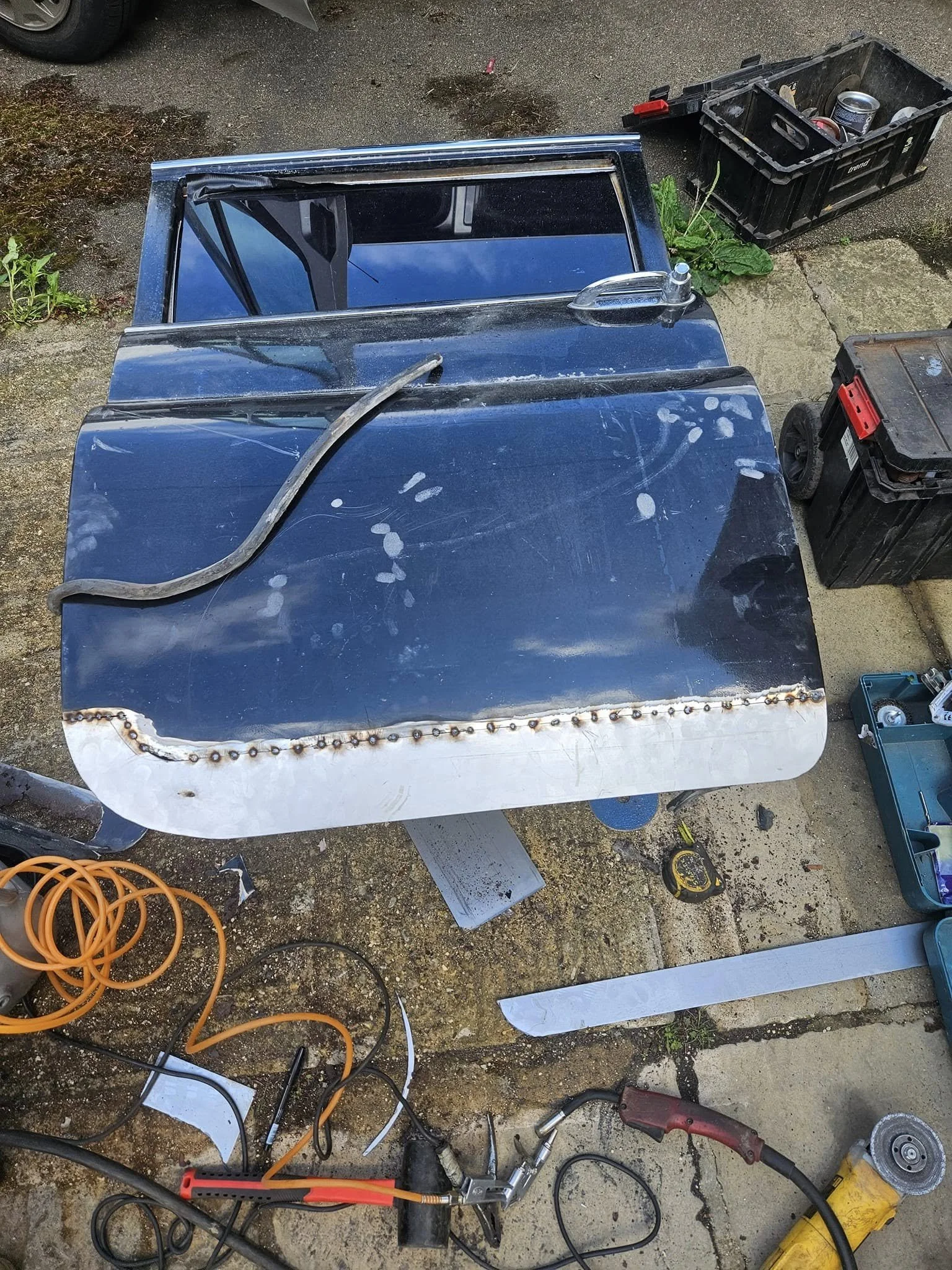 Metal car hood being welded, with tools and supplies around it on a concrete surface.