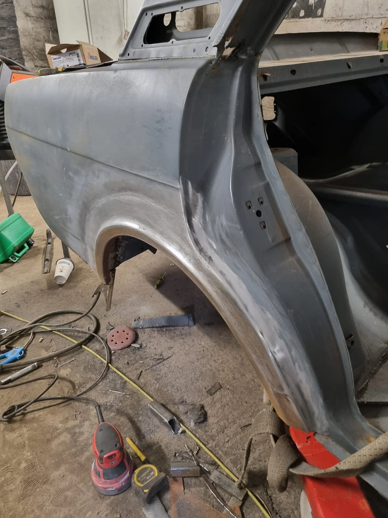 A car fender undergoing restoration or repair in a garage, with tools and equipment scattered on the floor.