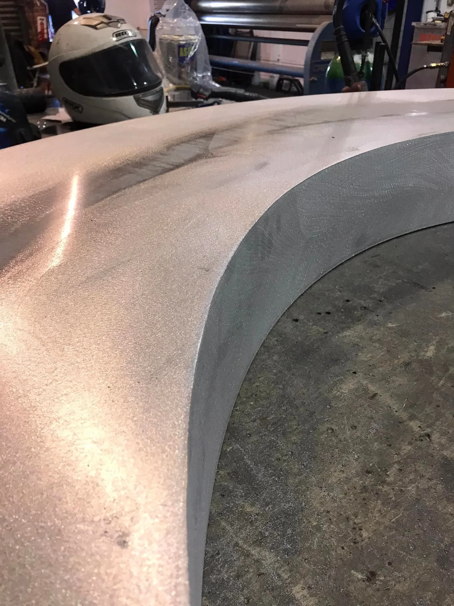 Close-up of a car's fender with a visible scratch or damage, metallic beige in color, with workshop tools and a helmet in the background.