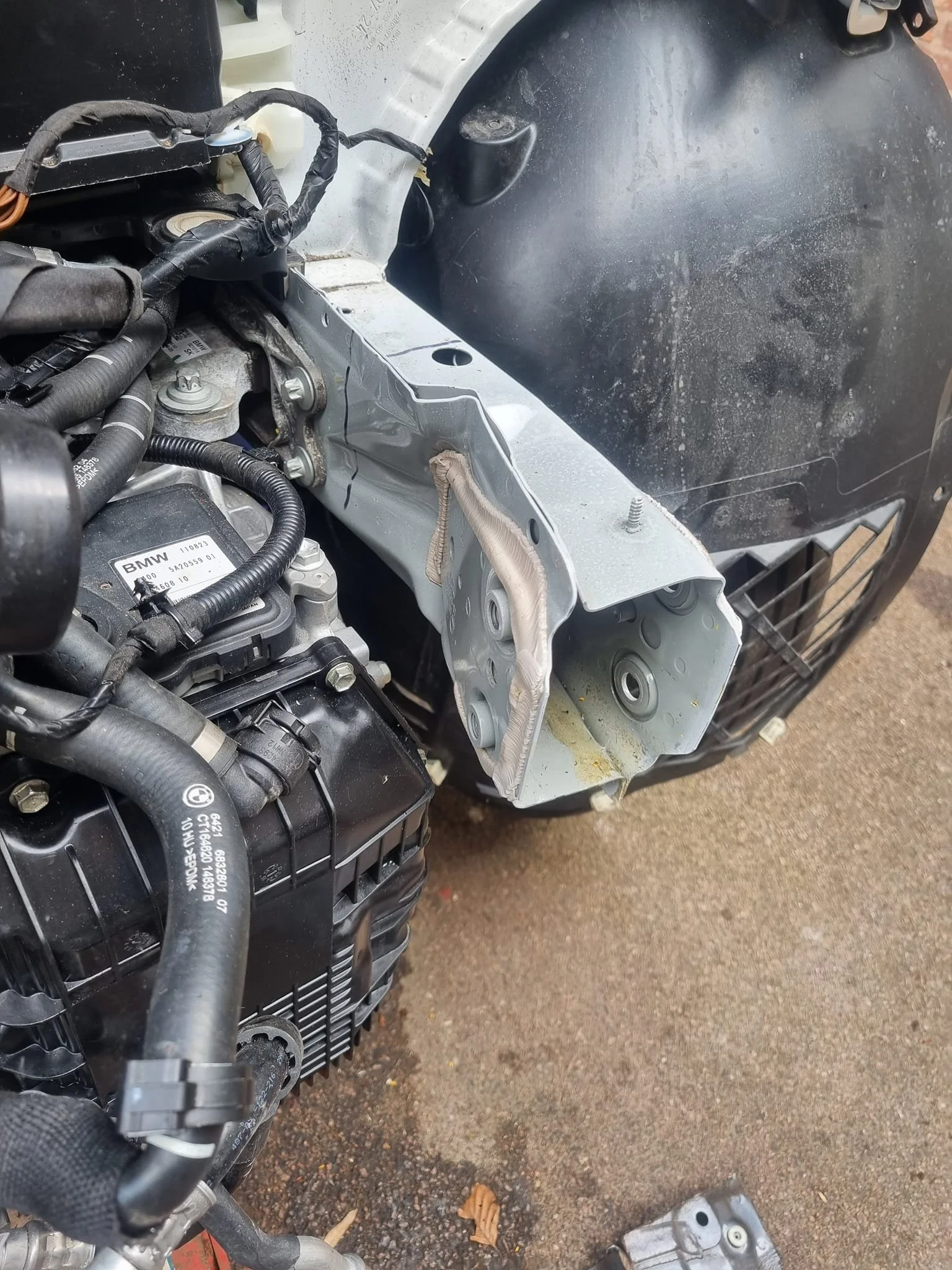 Engine bay of a vehicle with some parts removed, showing an exposed metal bracket, wiring, hoses, and components.
