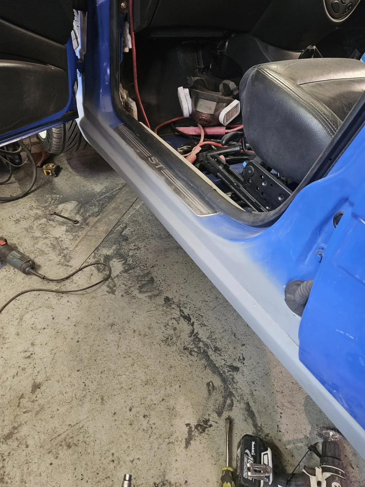 Inside a vehicle’s interior, showing the driver’s seat and leaning panel with red cables and a tool on rough, dirty garage floor.