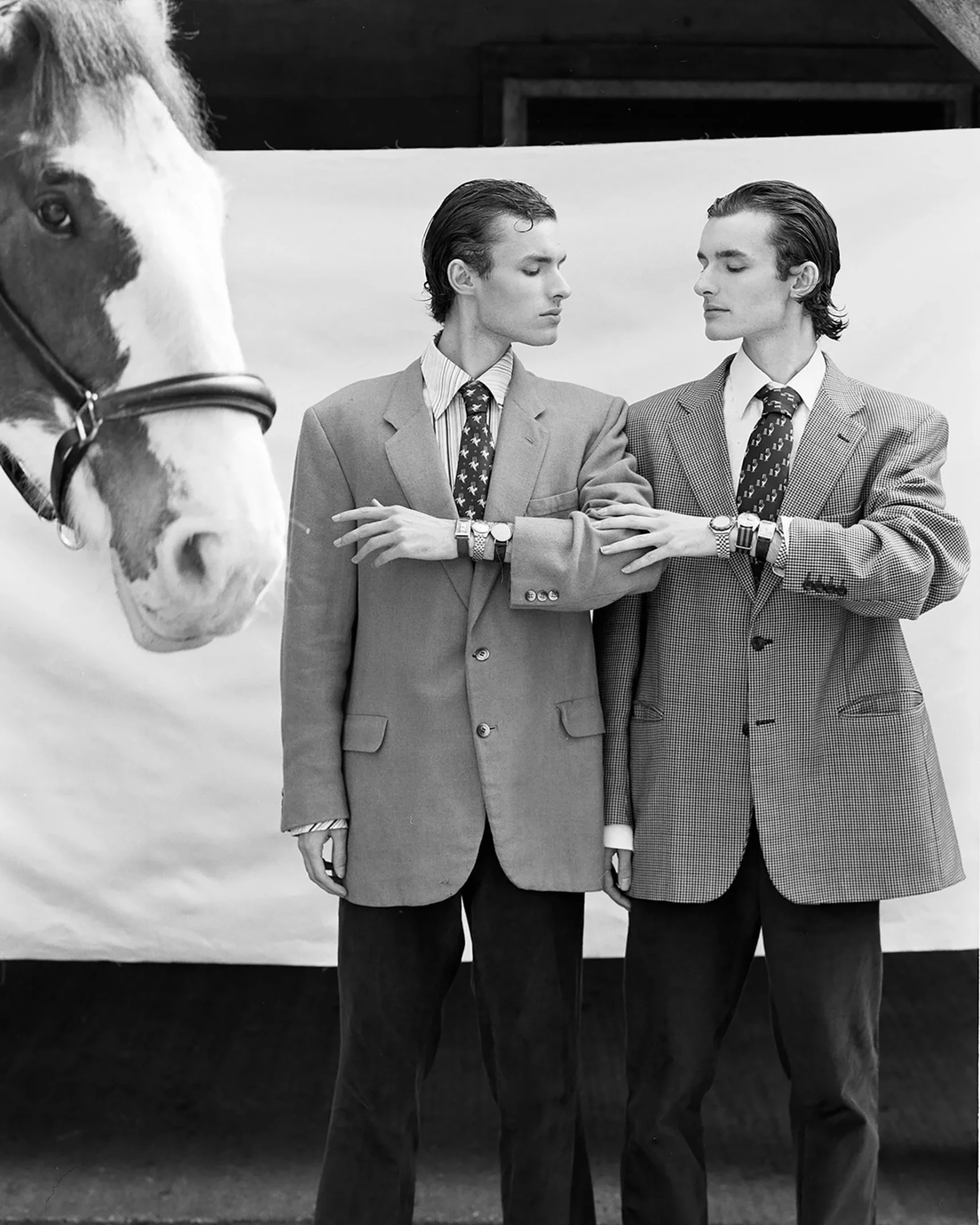 Two men in suits with watches and patterned ties are posed with their arms crossed, touching each other's arms, standing in front of a backdrop with a horse's face visible on the left side of the image. Surrealist photography.
