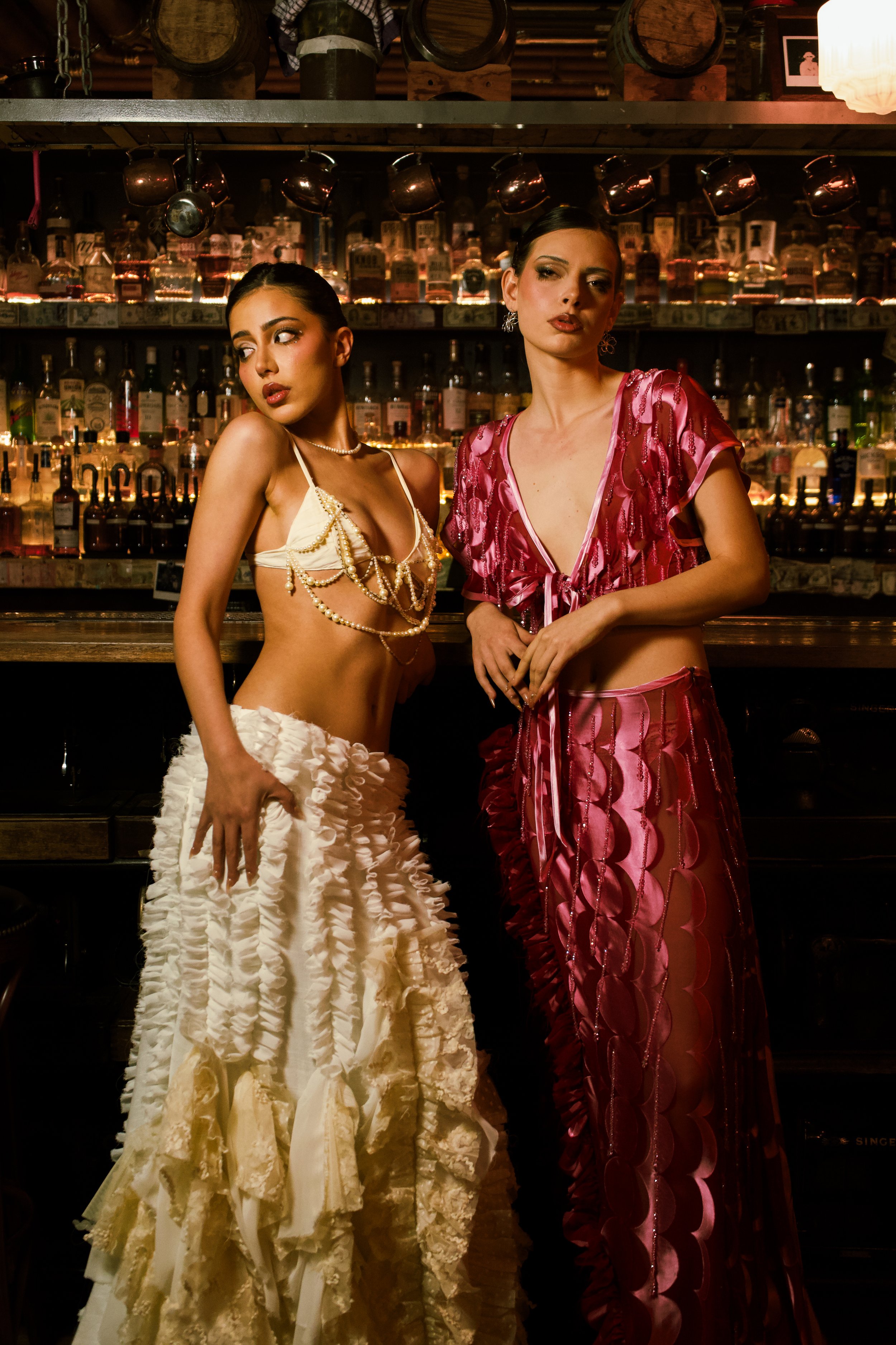 Two women in fancy attire posing in front of a bar with shelves of liquor bottles in the background.