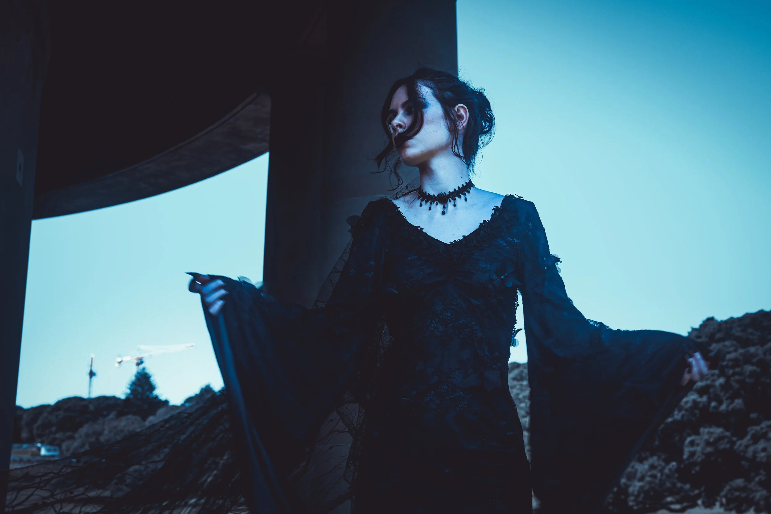 A woman dressed in black lace Gothic attire, standing outdoors with a modern bridge structure and cranes in the background during the evening or early night.