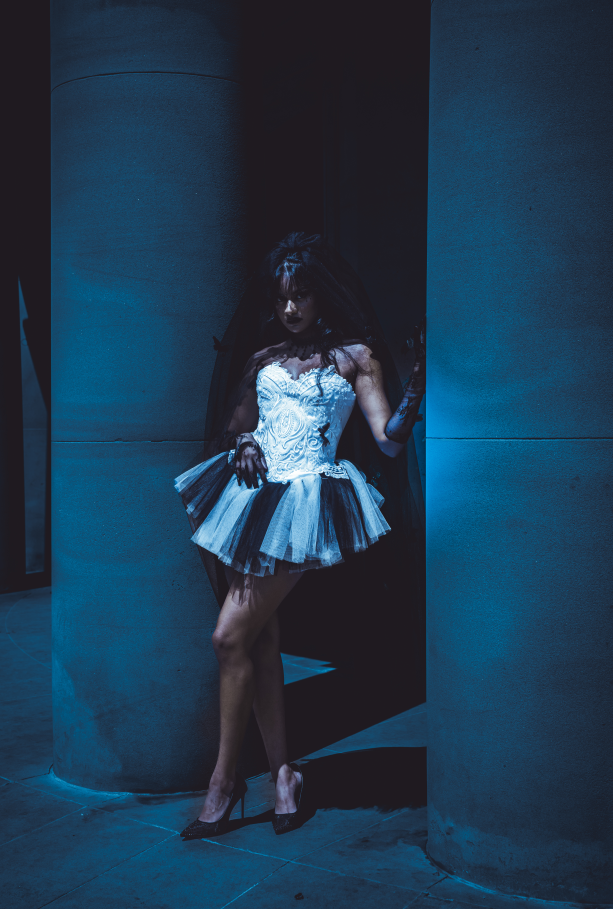 A woman in a white lace top and a black and white tutu with high heels standing in a dimly lit space with concrete pillars, partially framed in a doorway.