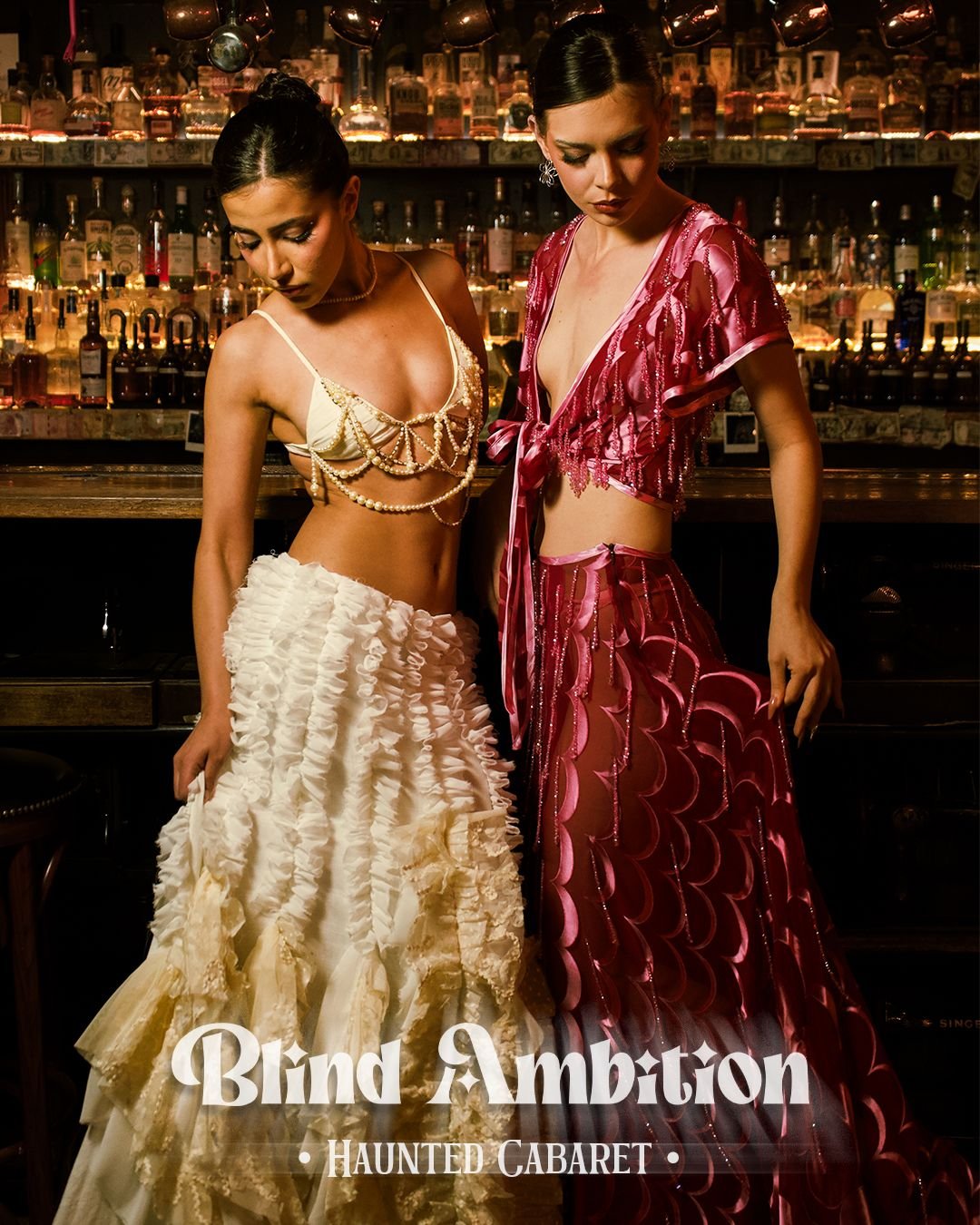 Two women dressed in elaborate, glamorous cabaret costumes pose in a dimly lit bar with a fully stocked liquor shelf in the background. One wears a cream-colored ruffled skirt and pearl necklace, while the other wears a pink sheer gown with scalloped patterns. The text 'Blind Ambition Haunted Cabaret' is overlaid on the image.