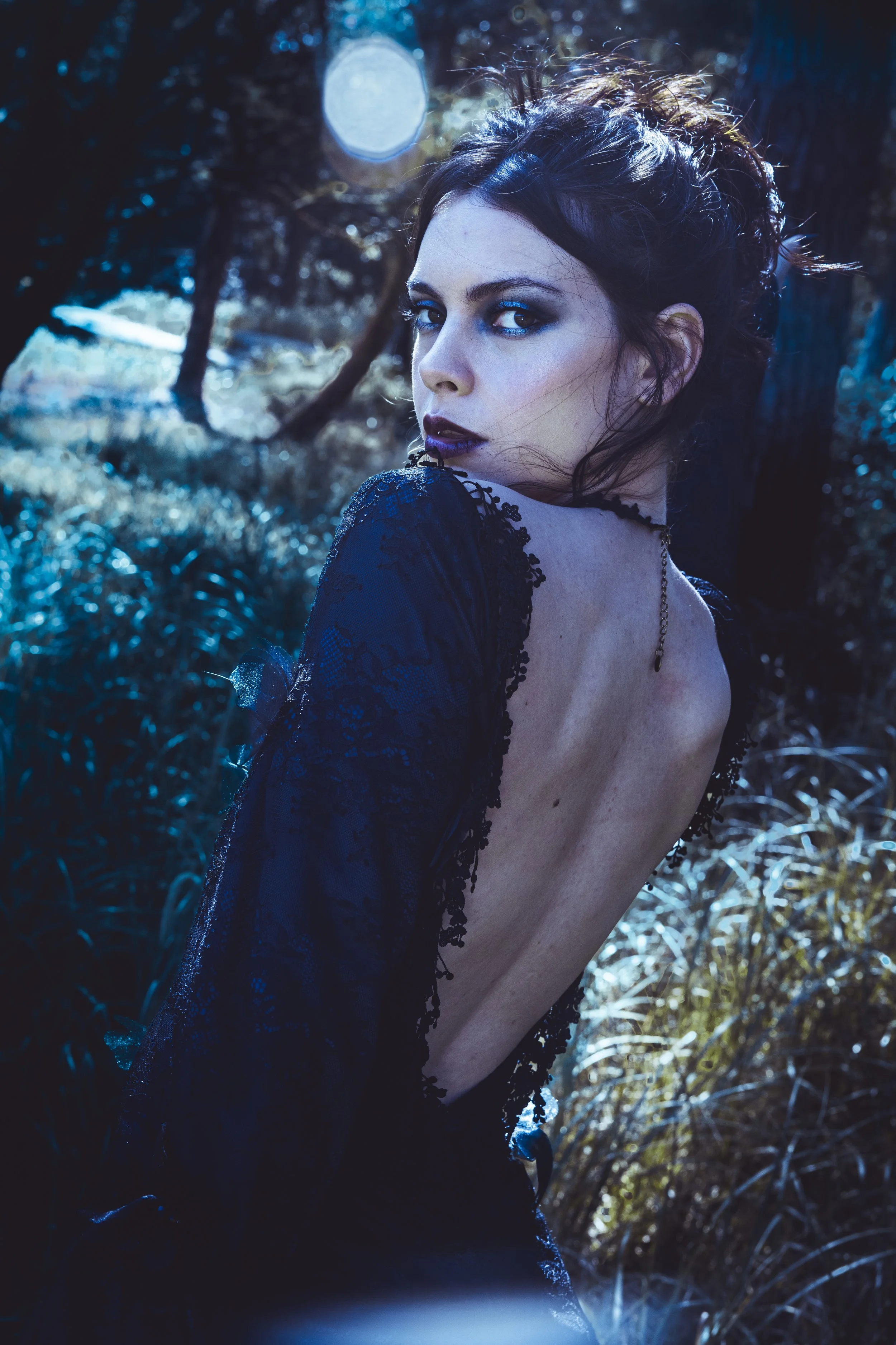 A woman with dark hair and dark makeup posing outdoors in a forested area with sunlight filtering through the trees, wearing a black lace dress with her back exposed.