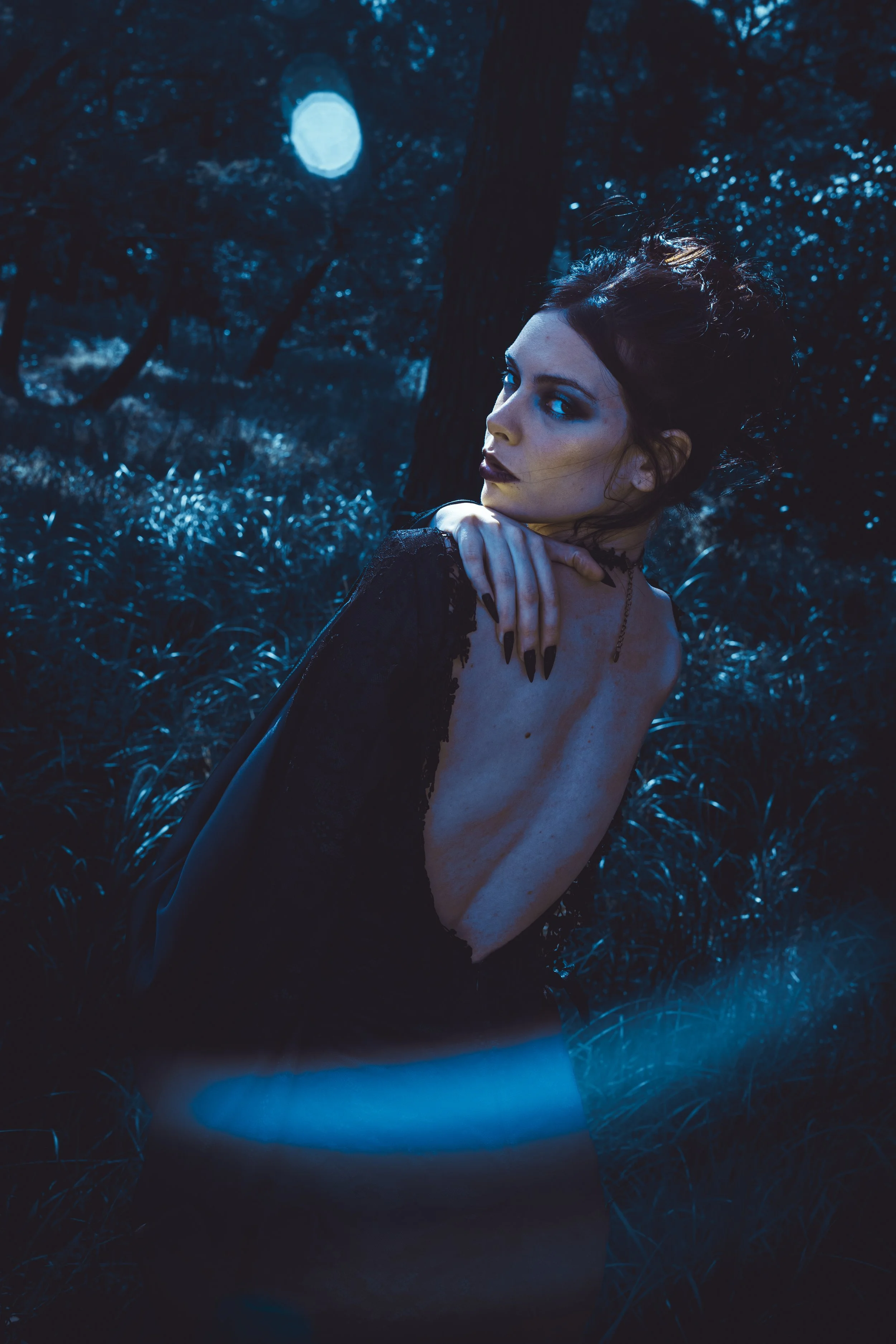 A woman with dark makeup and black nail polish posing in a dark, moonlit forest with blue lighting.