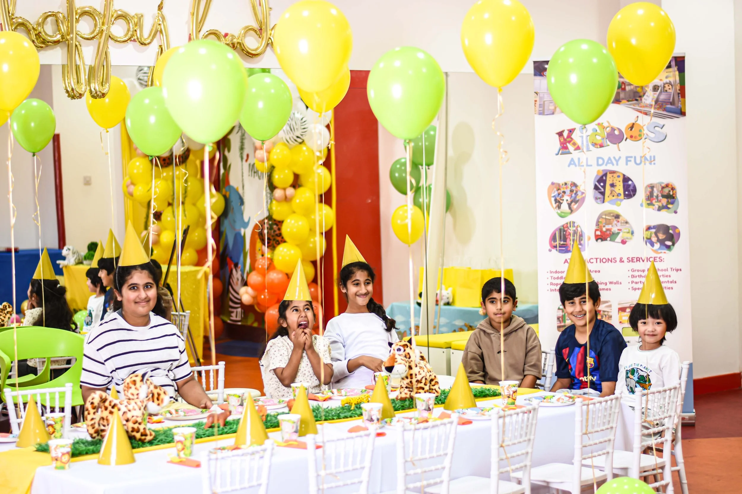 Children wearing yellow party hats sitting at a decorated birthday party table with animal-themed decorations, balloons, and a colorful backdrop.