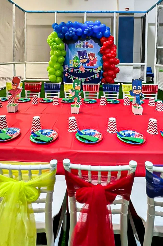 Colorful superhero themed birthday party setup with a table decorated with plates, cups, and cutouts of superhero characters, a backdrop with balloons in red, blue, green, and purple, and a banner that says "Happy Birthday."