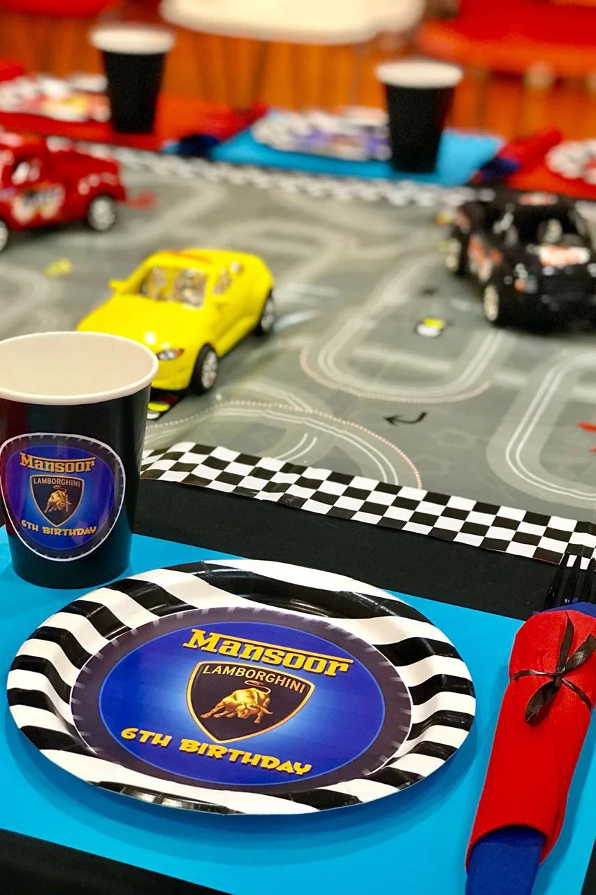 A children's birthday party table with a racing car theme, featuring a plate and cup with Lamborghini logos and the words '6th Birthday.' There are toy cars on a racing track mat in the background, along with a red rolled-up napkin on the table.