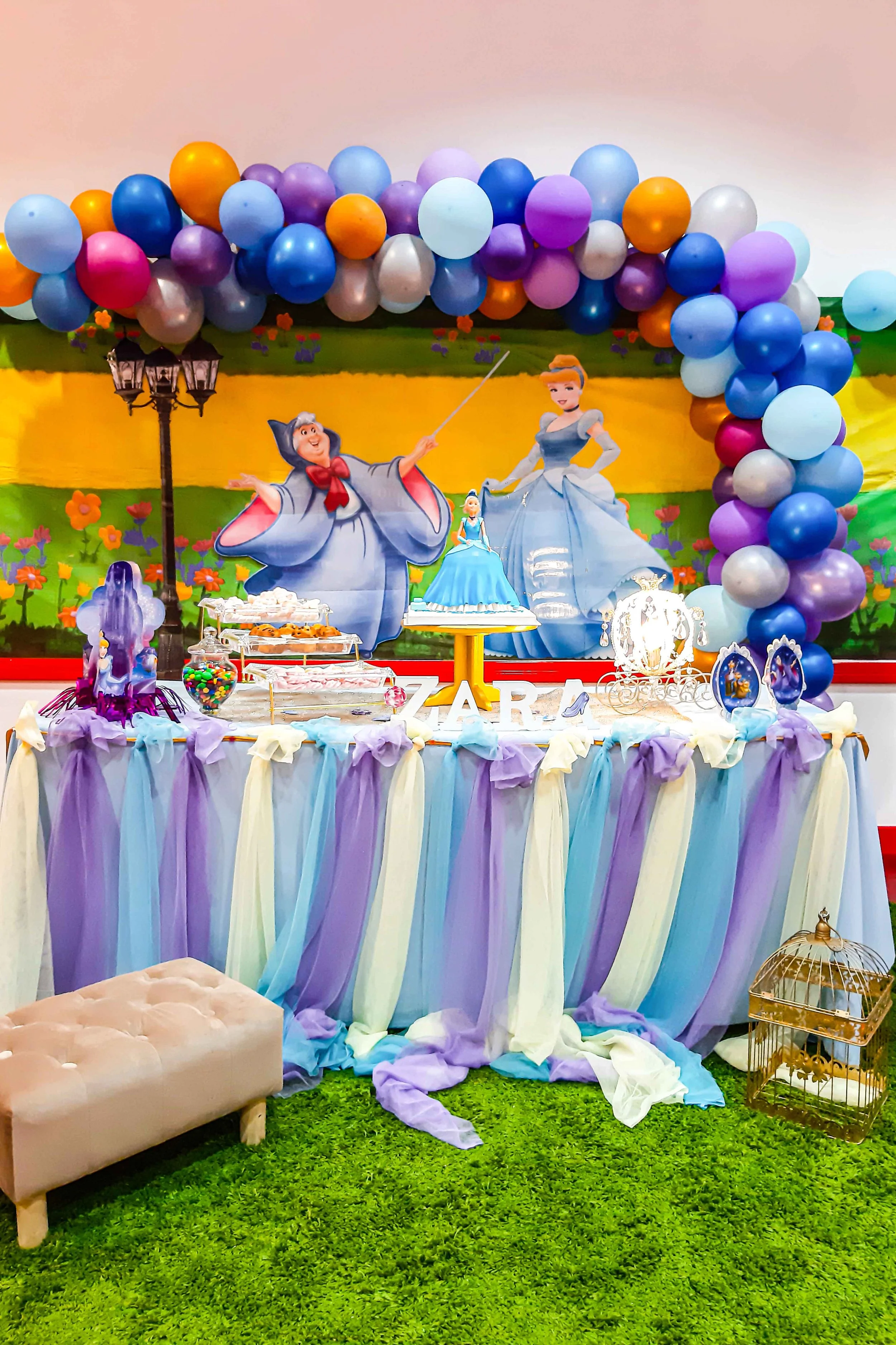 A Disney princess-themed birthday party table with purple, blue, and white decorations, featuring a Cinderella backdrop with balloon arch, princess figurines, and a cake.
