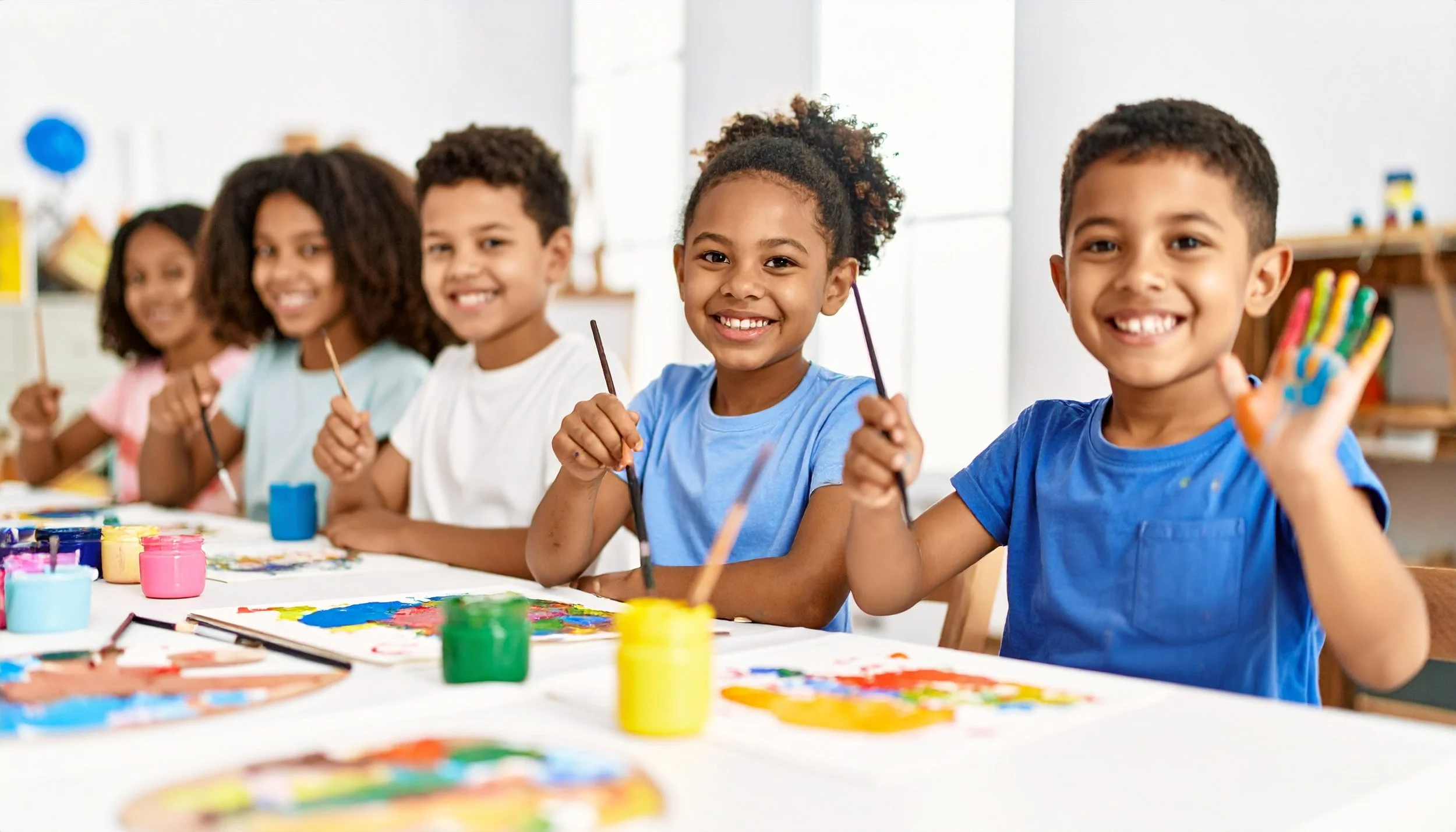 Five children sitting at a table, smiling, and holding paintbrushes during a painting activity in a classroom.