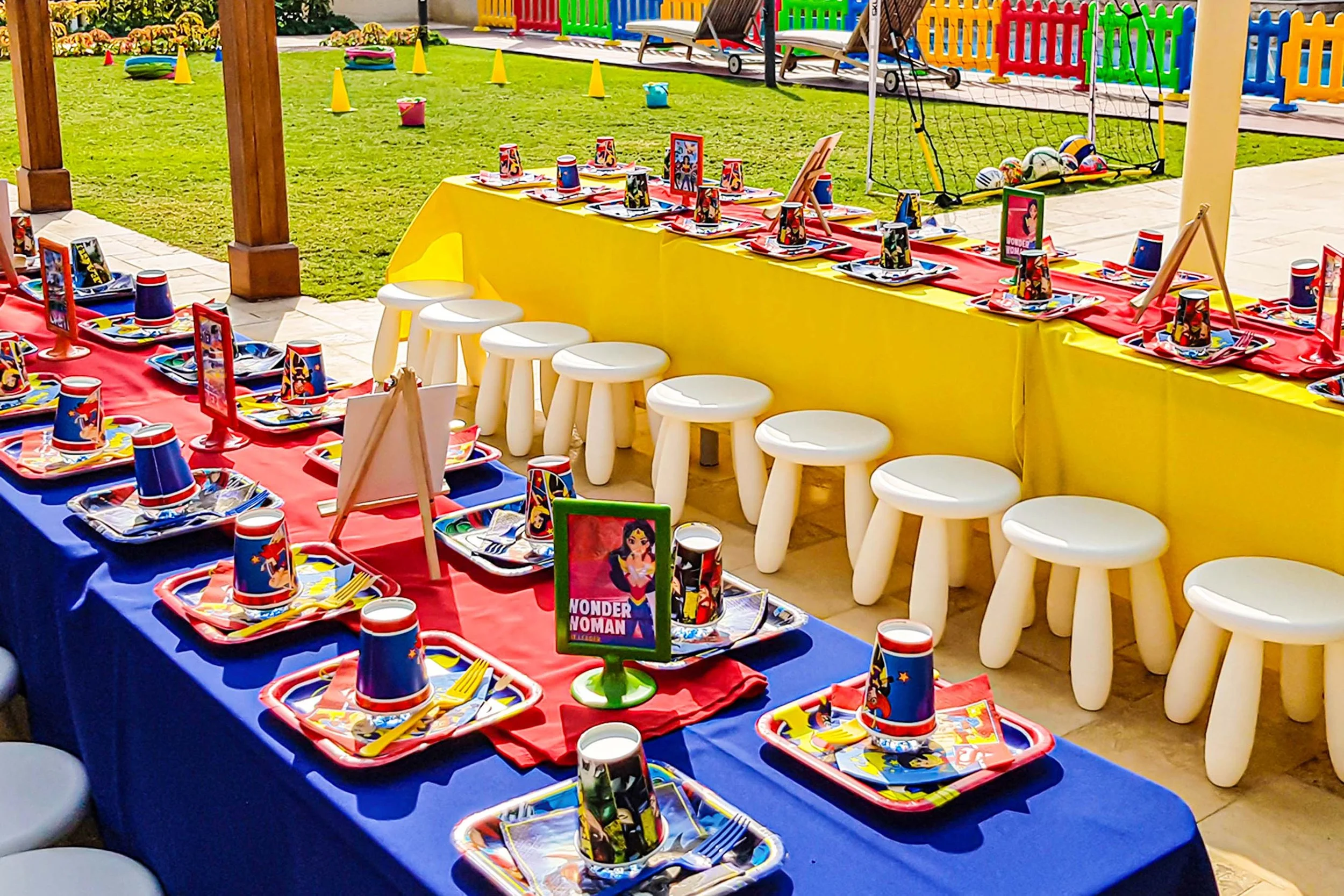 Colorful outdoor birthday party setup with a table decorated in red, blue, and yellow themed plates, cups, and napkins featuring Wonder Woman images. Small white stools are arranged along the table, with a yellow and red banner of Wonder Woman on the