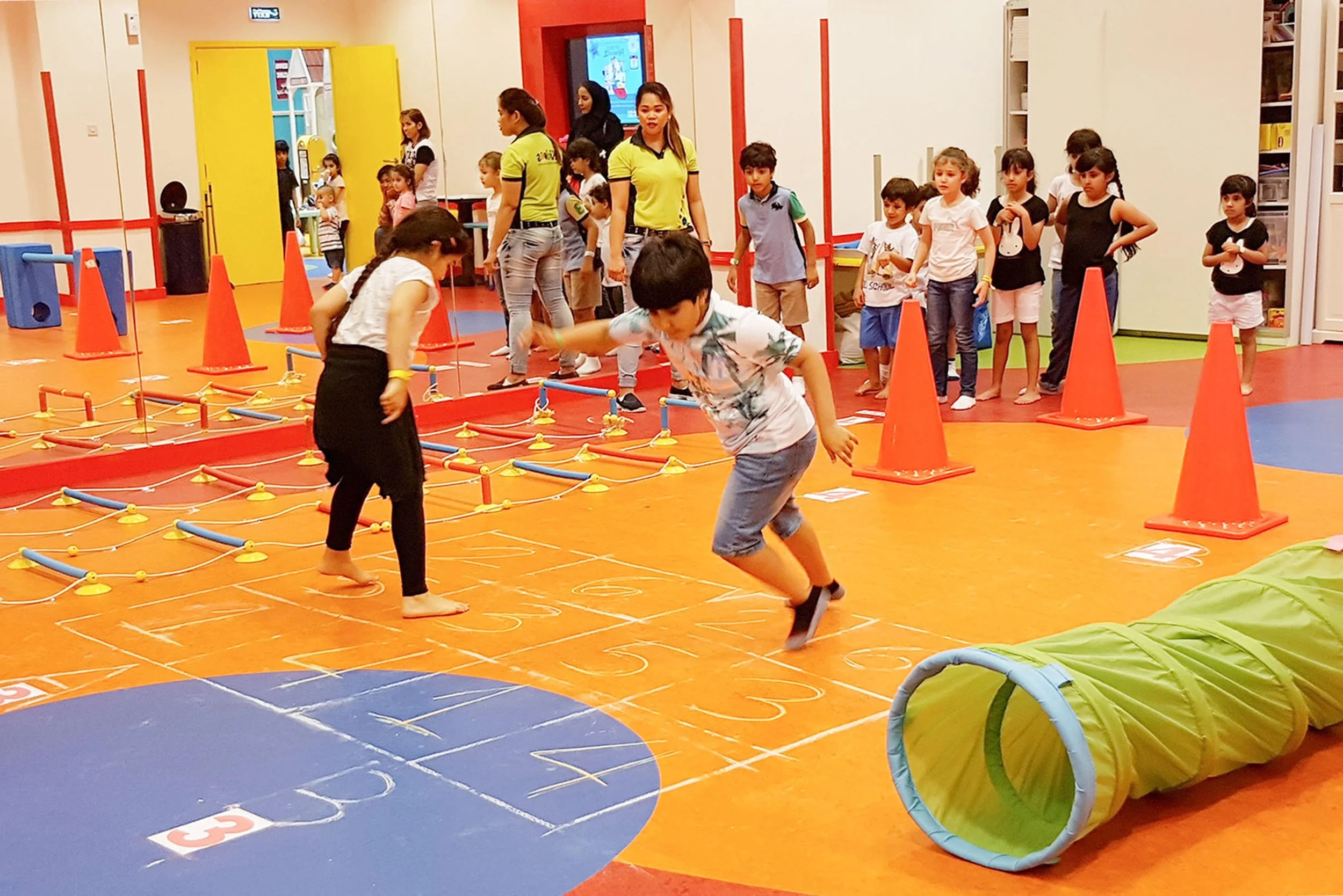 Children playing and jumping in an indoor play area with orange cones, ladders, and colorful floor markings, surrounded by other kids and adults.