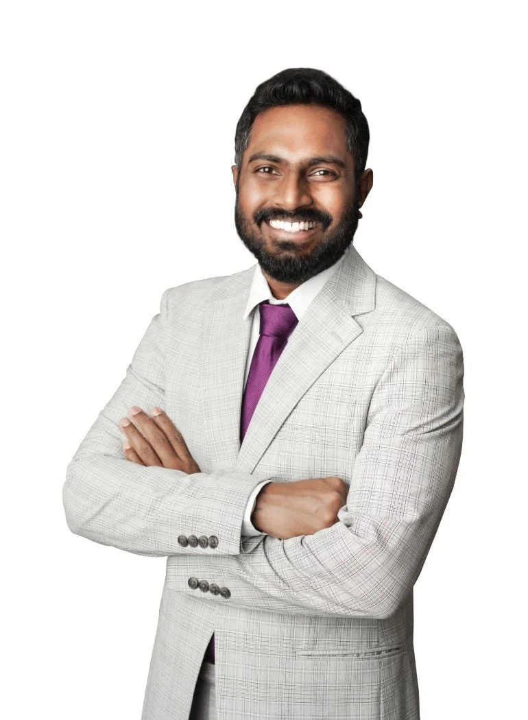 A man with dark hair and a beard smiling, wearing a light gray suit, white shirt, and purple tie, standing with arms crossed against a plain white background.