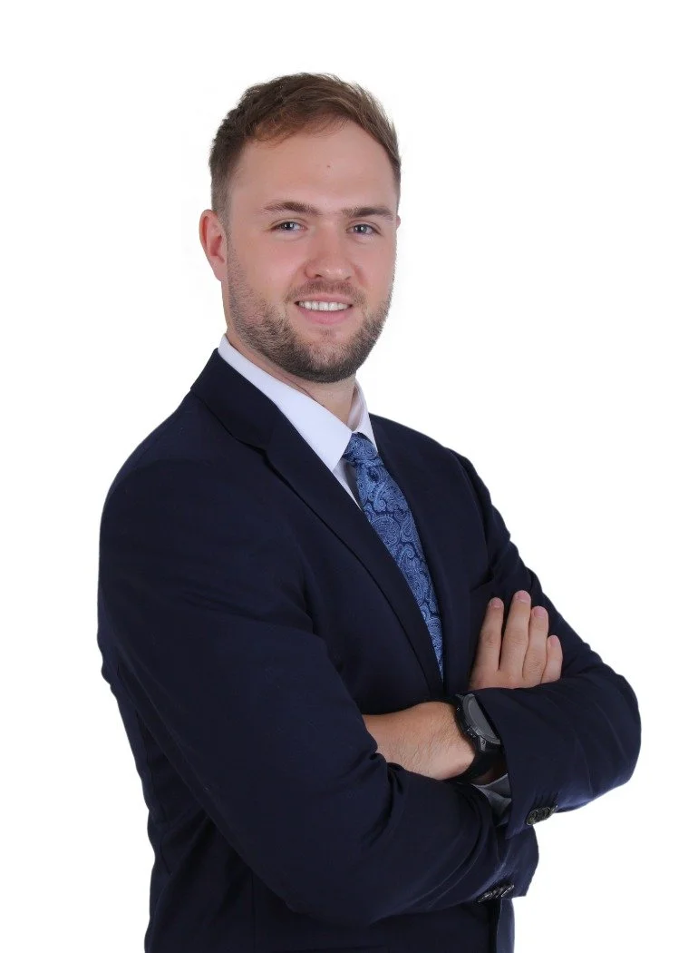A young man with light brown hair and a beard, wearing a dark suit, white shirt, and patterned blue tie, standing with arms crossed and smiling at the camera against a white background.