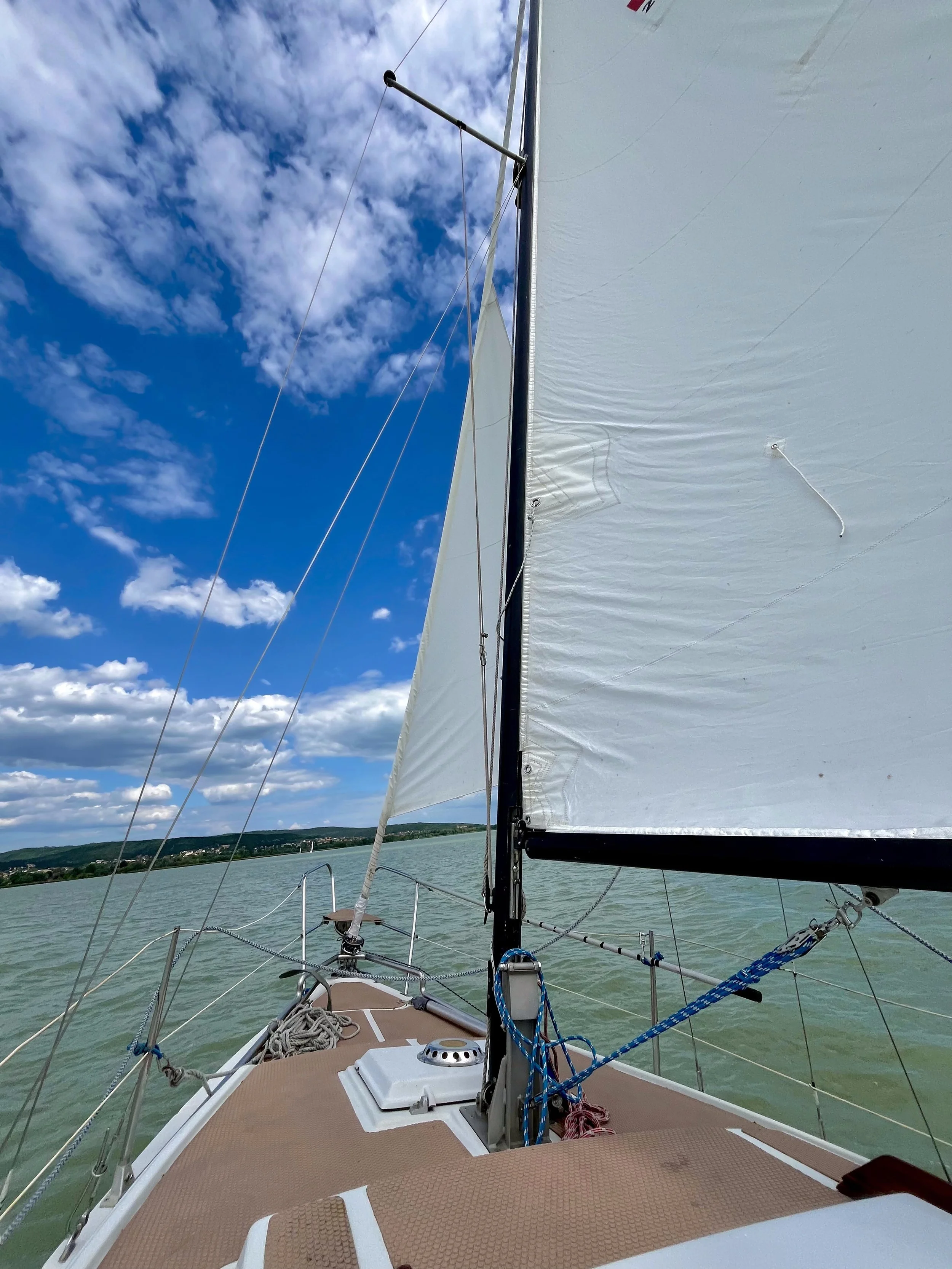 View from the bow of a sailboat on a lake, showing the deck, sail, rigging, and blue sky with clouds in the background.