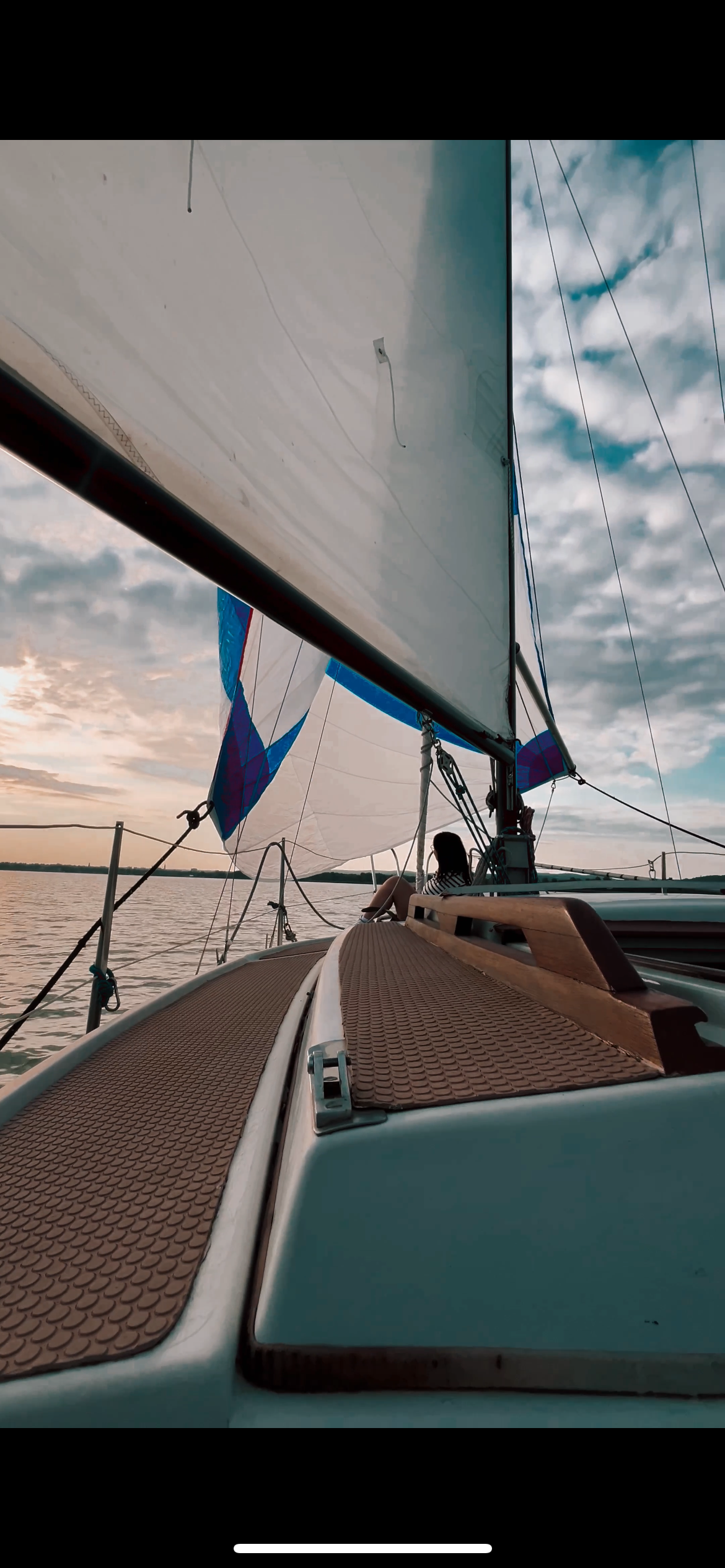 View from the bow of a sailboat on the water during sunset, showing sails, ropes, and a woman sitting at the back of the boat.