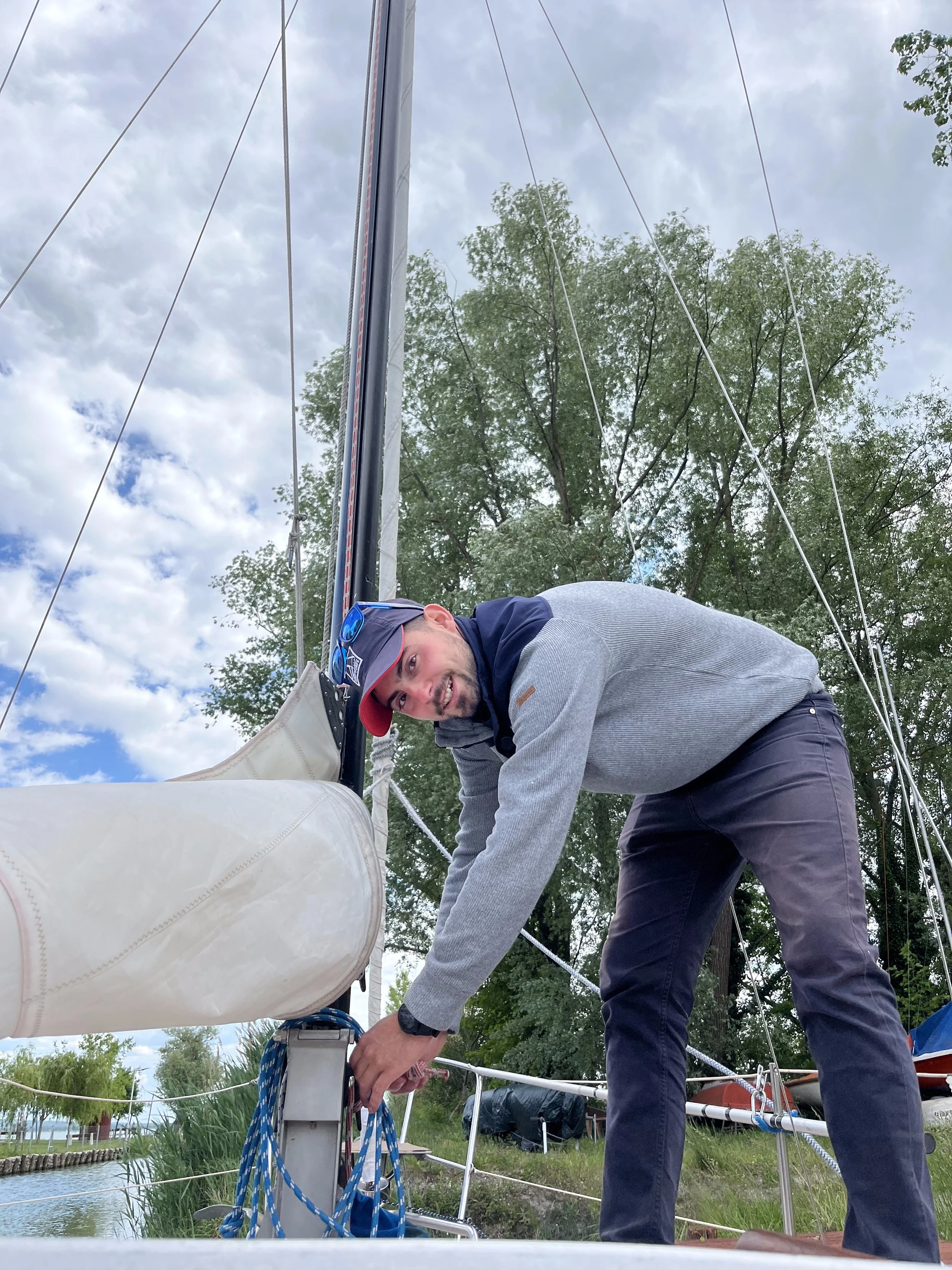 Man tying ropes on a sailboat docked by a lake, wearing a gray and navy jacket, navy pants, and a baseball cap, with green trees and cloudy sky in the background.
