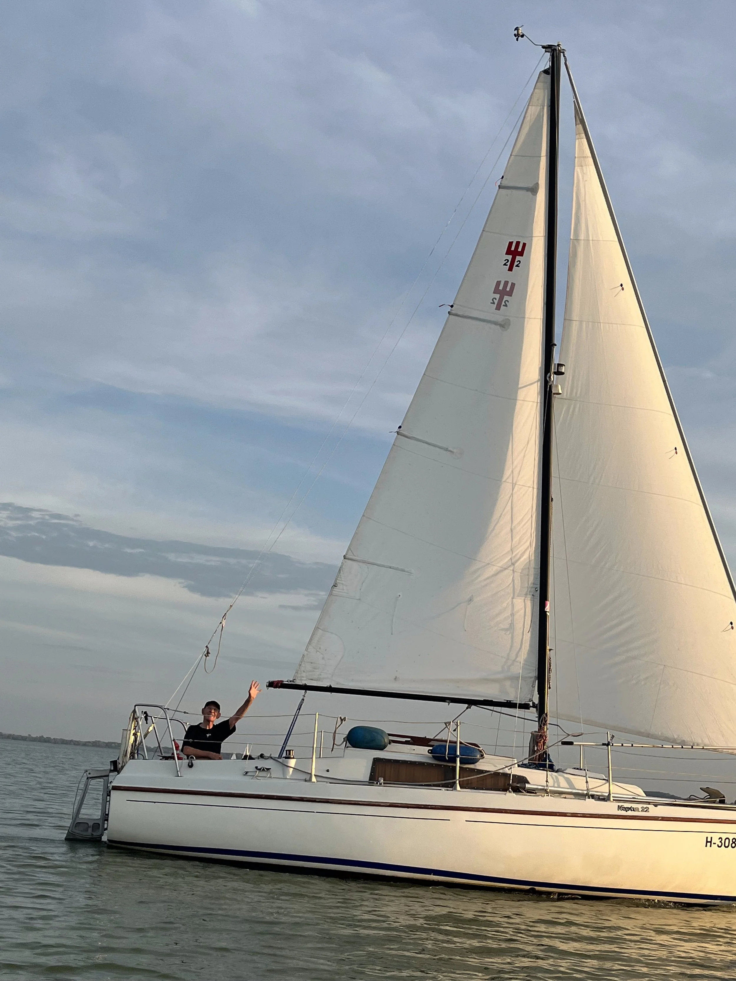 Person waving while sailing on a white sailboat with beige sails on calm water during daytime.