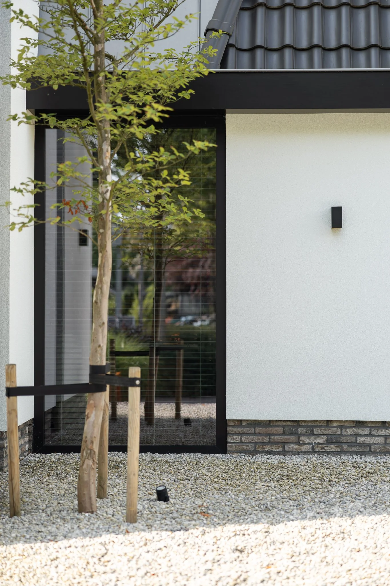 Glass entrance connecting two sections of a white villa beside a young tree and gravel garden