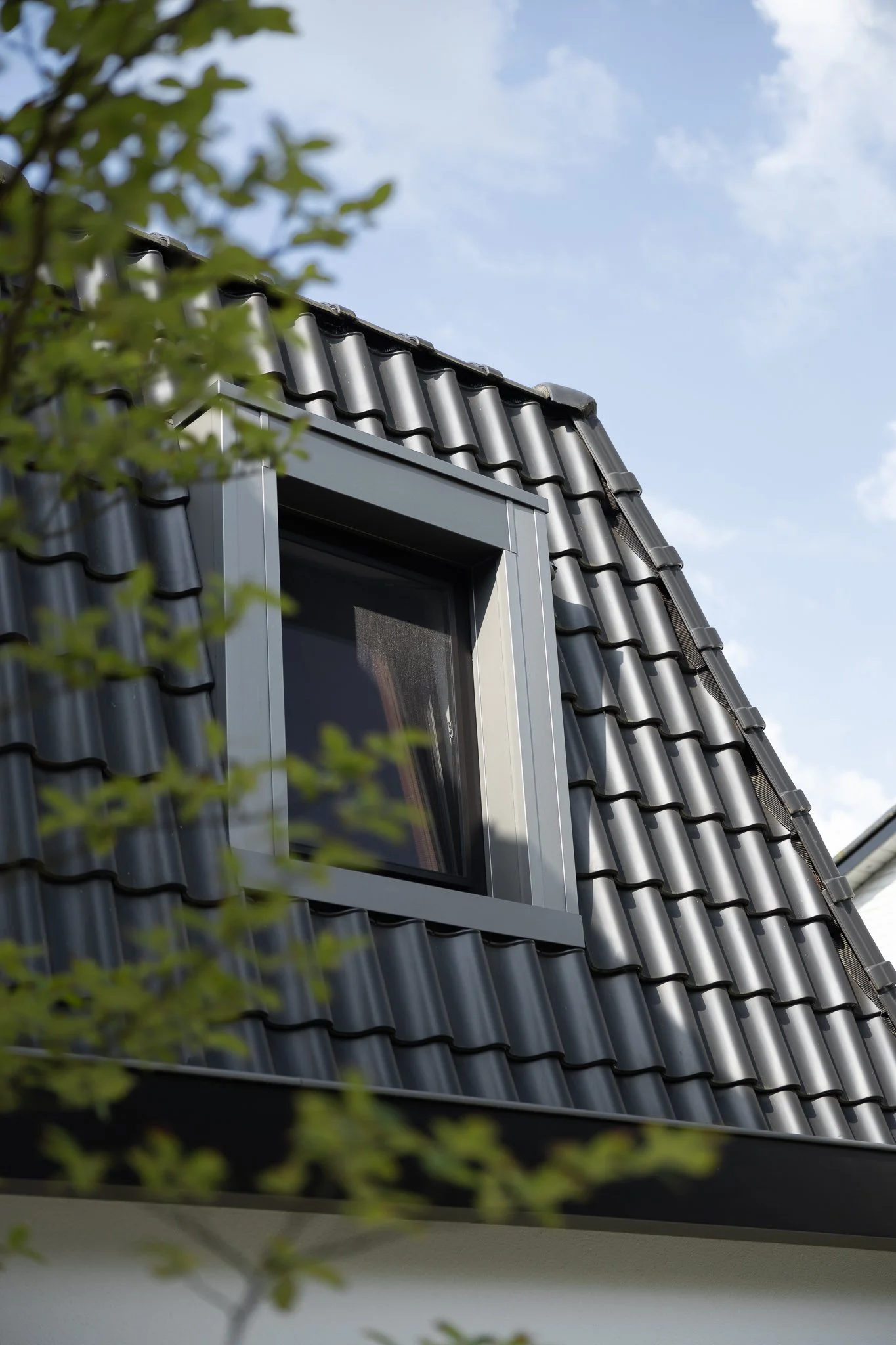 Modern dormer window integrated into a dark tiled roof and framed by green branches