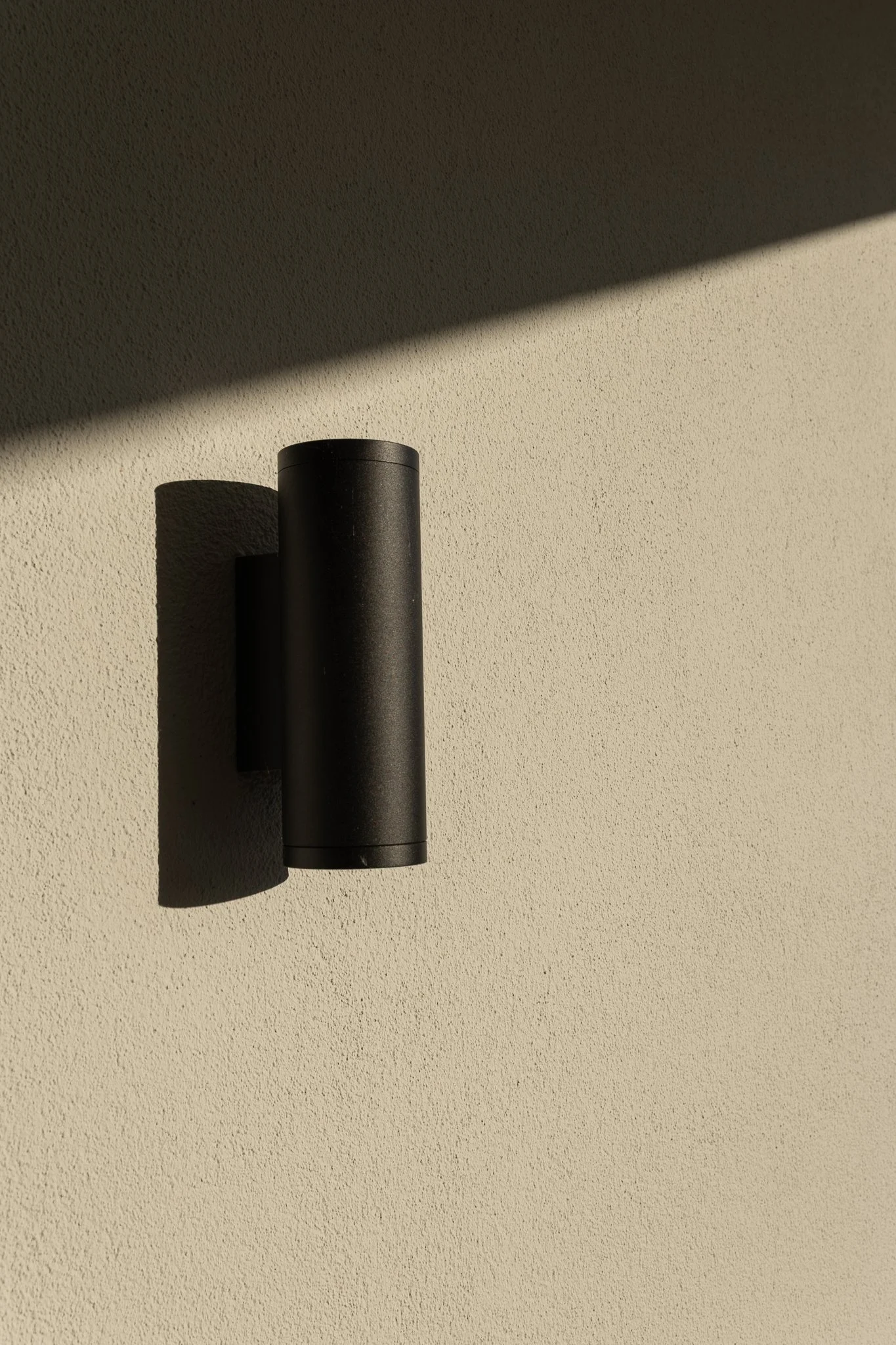 Minimal black outdoor wall light casting a strong shadow on a textured beige façade